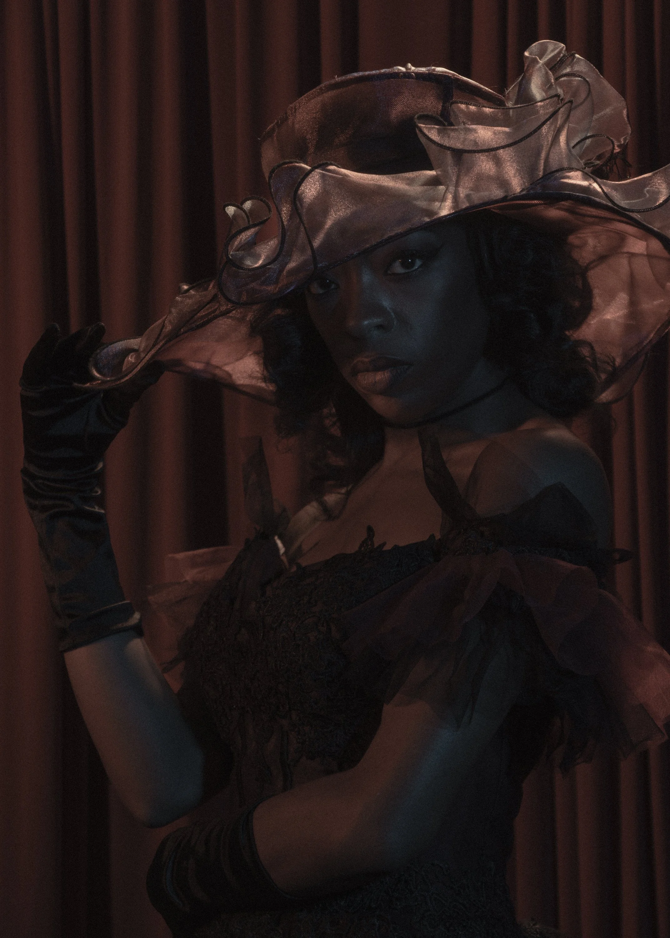 A woman wearing a large, translucent, dark-colored hat with ruffled edges, black gloves, a black lace dress, and long striped stockings, posing in front of a dark wood-paneled wall.