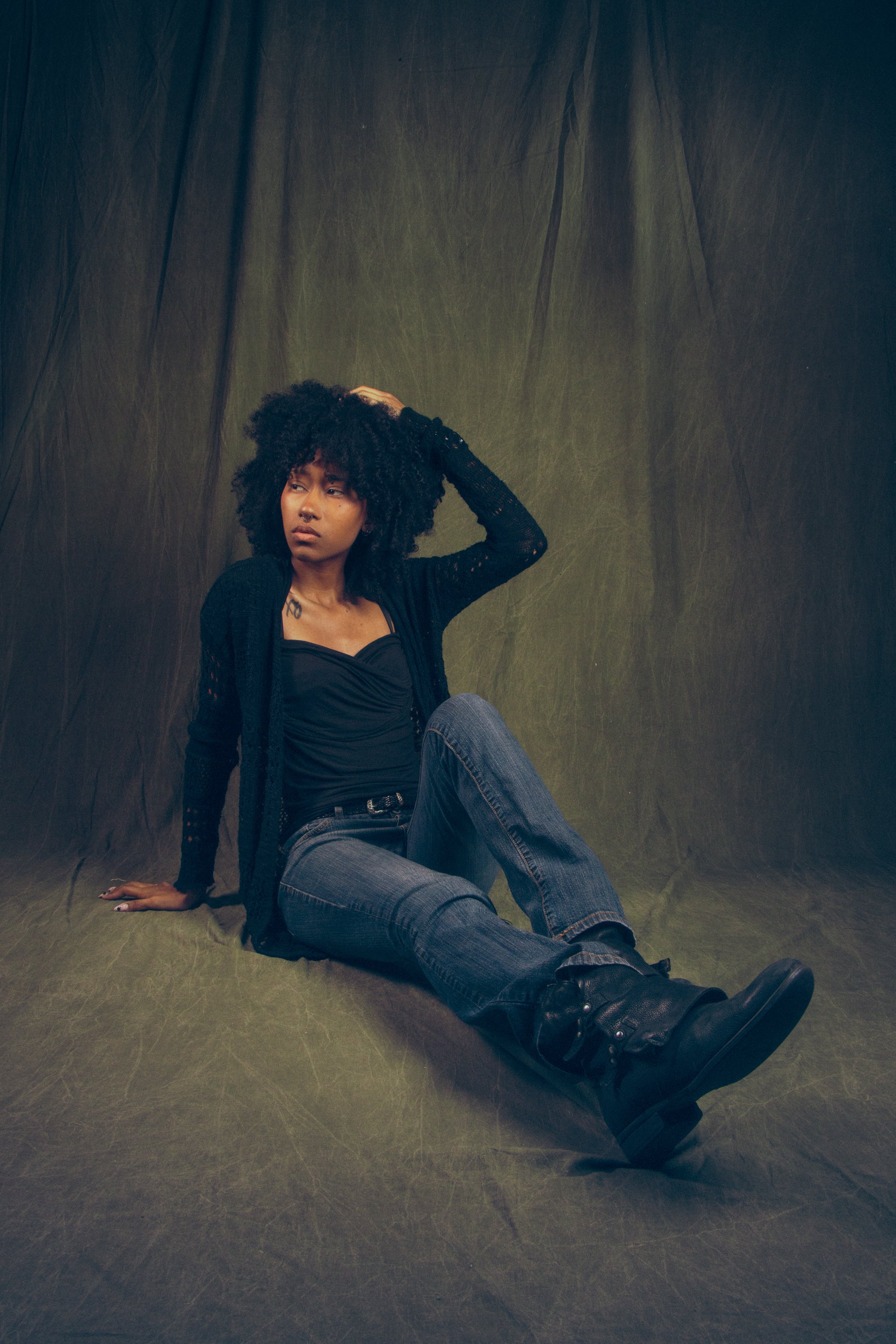 A young woman with curly hair and a nose piercing sitting on the floor against a dark backdrop, wearing a black top with a cardigan, jeans, and black boots. She looks thoughtfully into the distance.