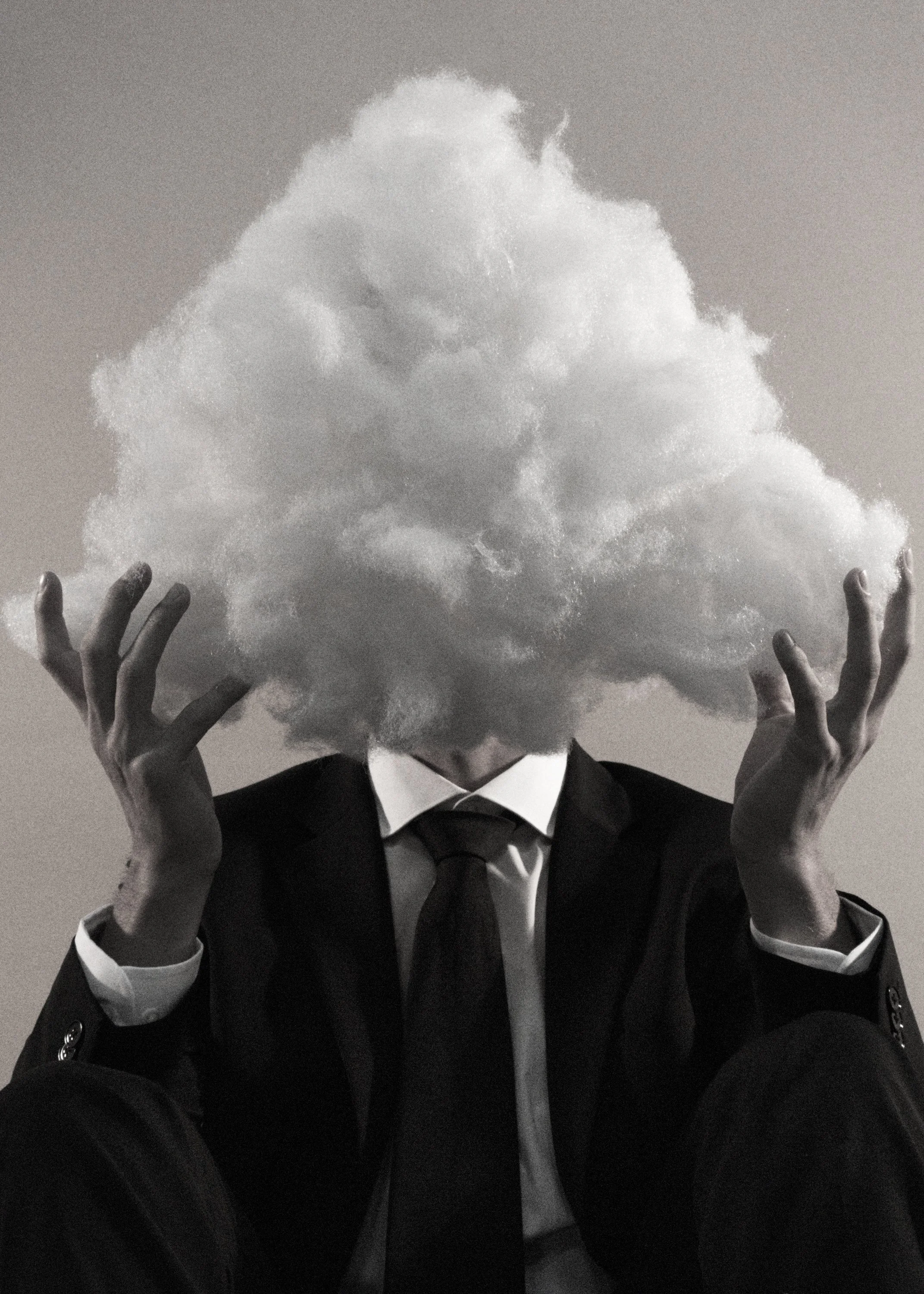 A person in a suit and tie with their head replaced by a cloud, sitting with legs crossed and hands raised slightly.