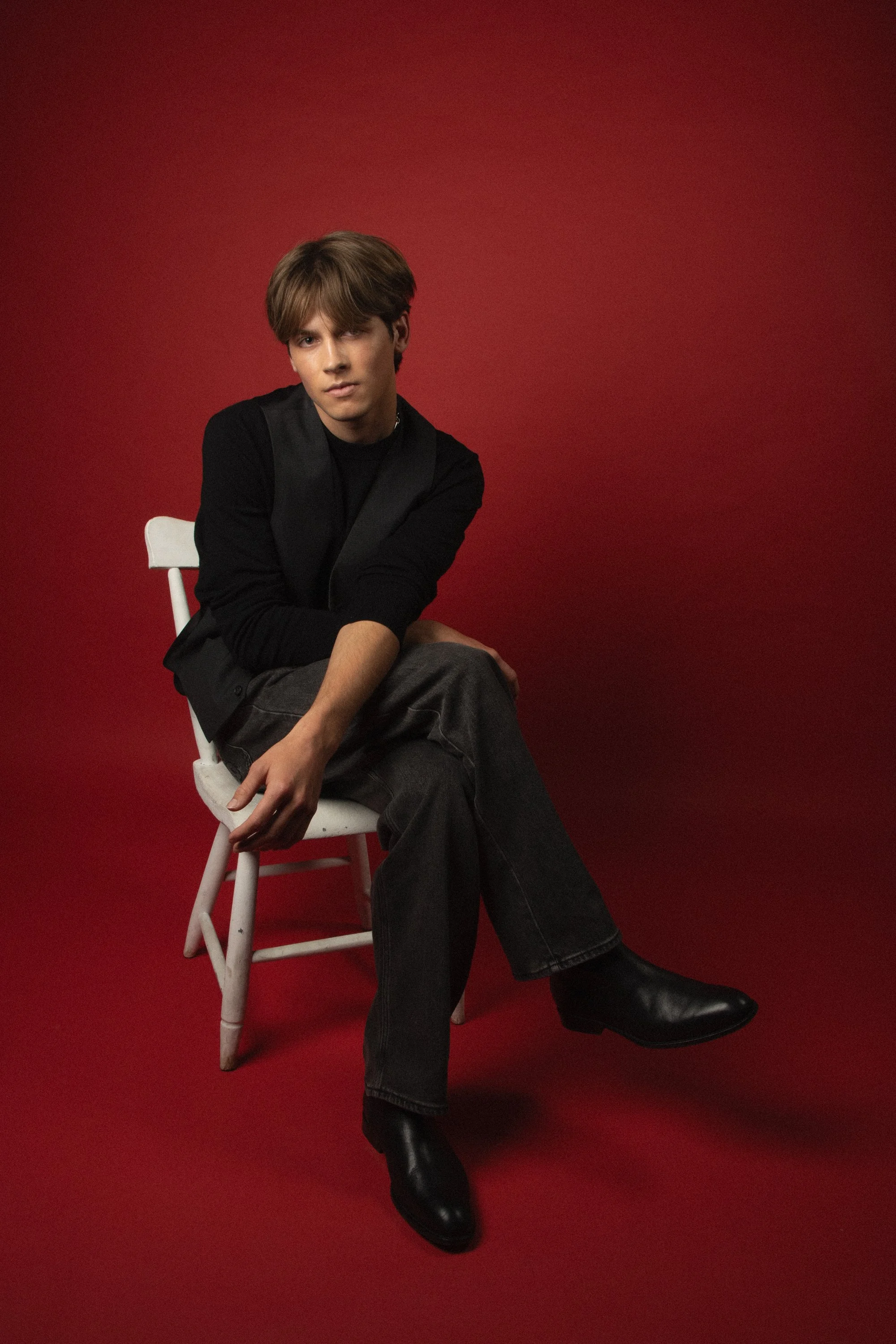 A young man with brown hair sitting on a white wooden chair against a red background. He is dressed in a black jacket, black boots, and dark pants, looking directly at the camera with a serious expression.