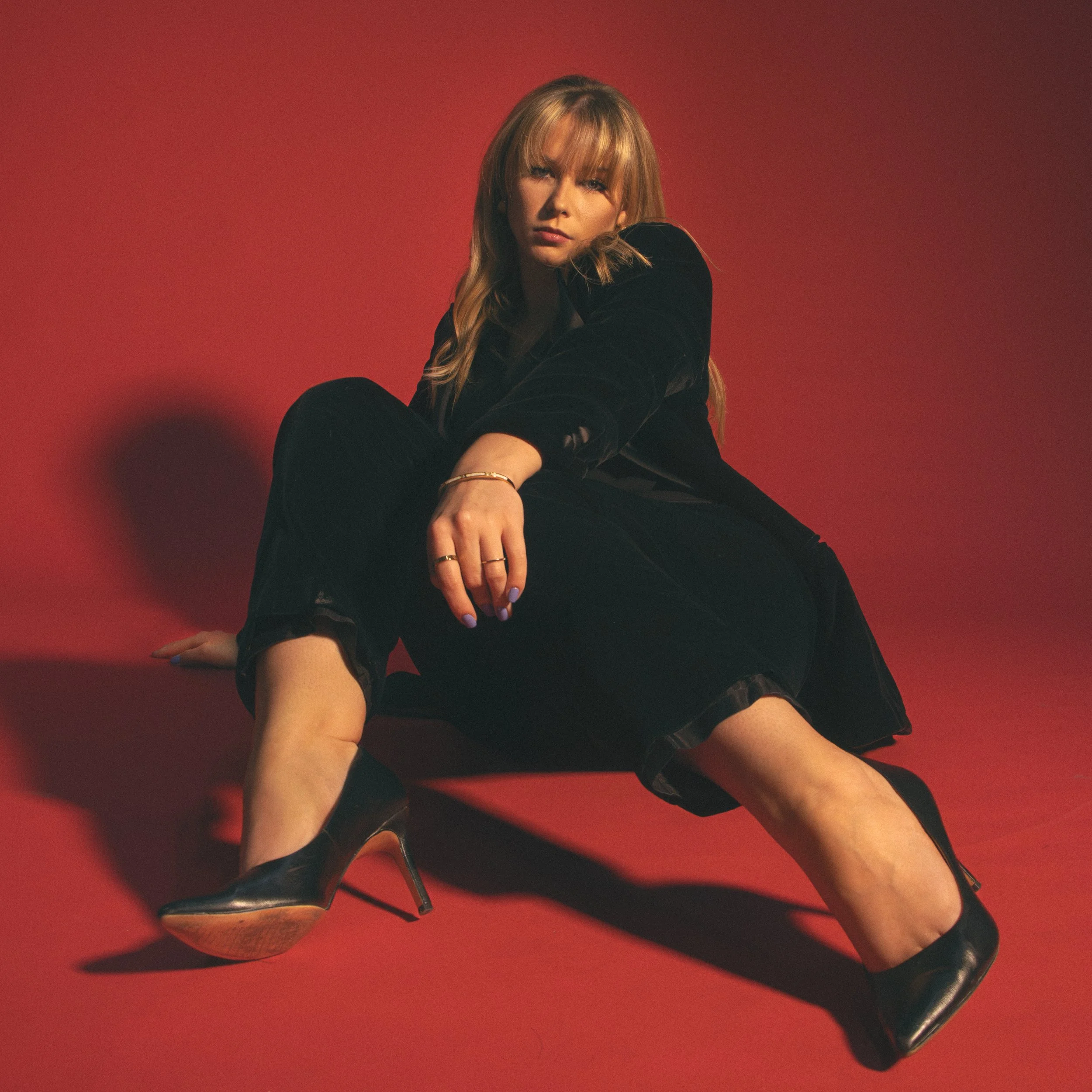 A woman with blonde hair wearing black clothing sitting on the floor against a red background, dressed in black high heels, with her arm resting on her knee.