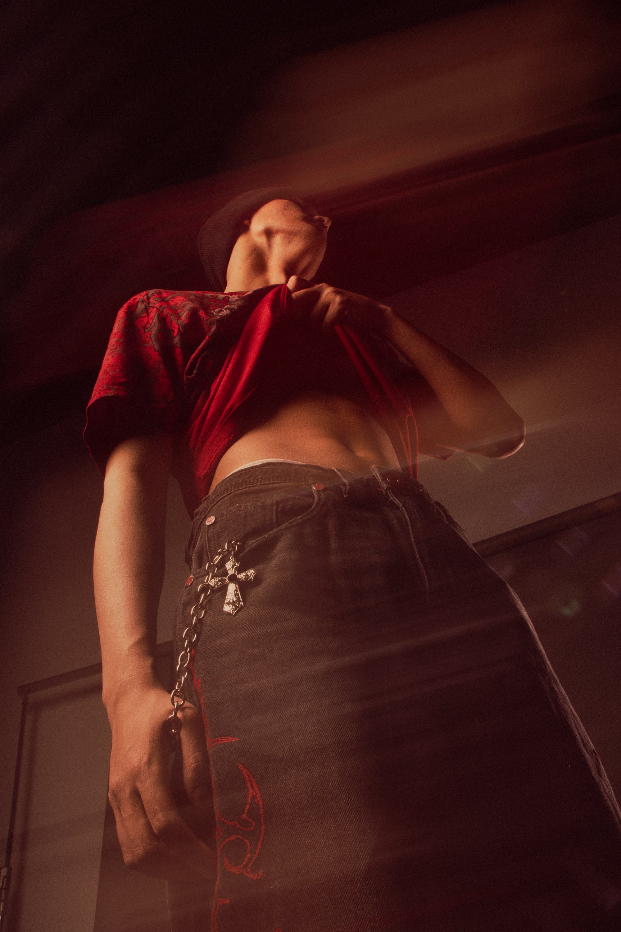 Low angle photo of a person wearing a black beanie, red and black graphic t-shirt, and black pants with a chain accessory, standing in a dimly lit room.