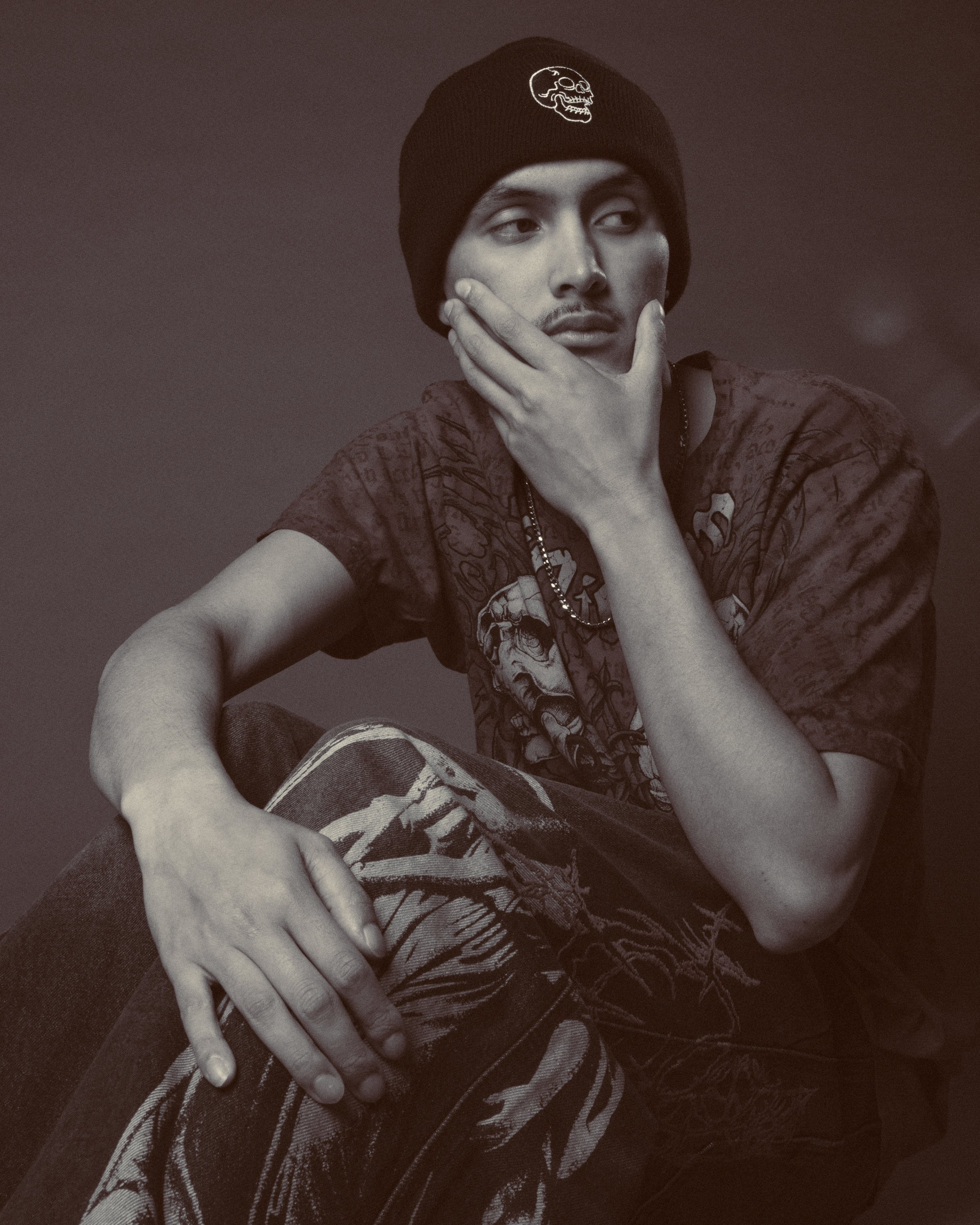 Young man wearing a black beanie with a skull logo, sitting with one arm resting and the other hand on his chin, wearing a graphic t-shirt and patterned pants.