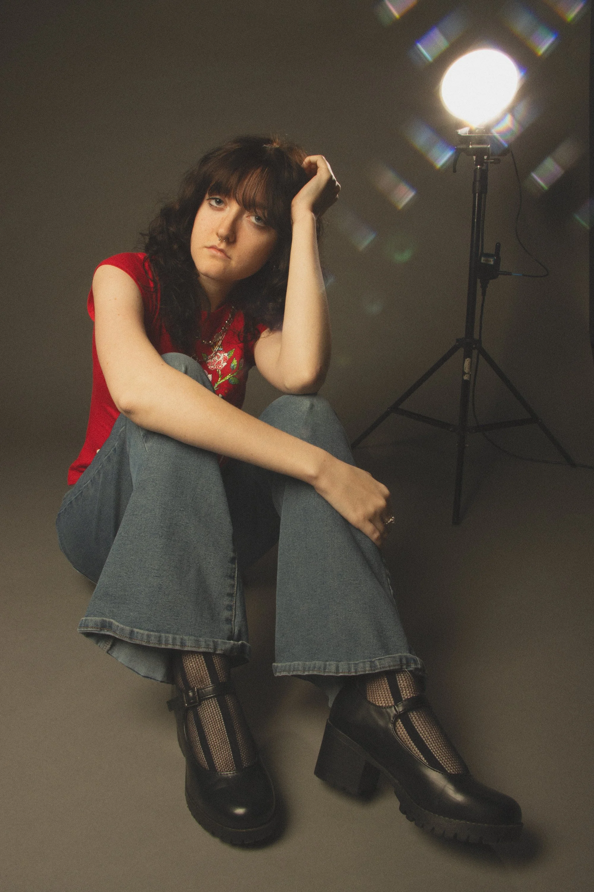 A person sitting on the floor with crossed legs, wearing a red T-shirt with colorful embroidery, ripped jeans, patterned fishnet stockings, and black boots, in a studio with a single spotlight to their right.
