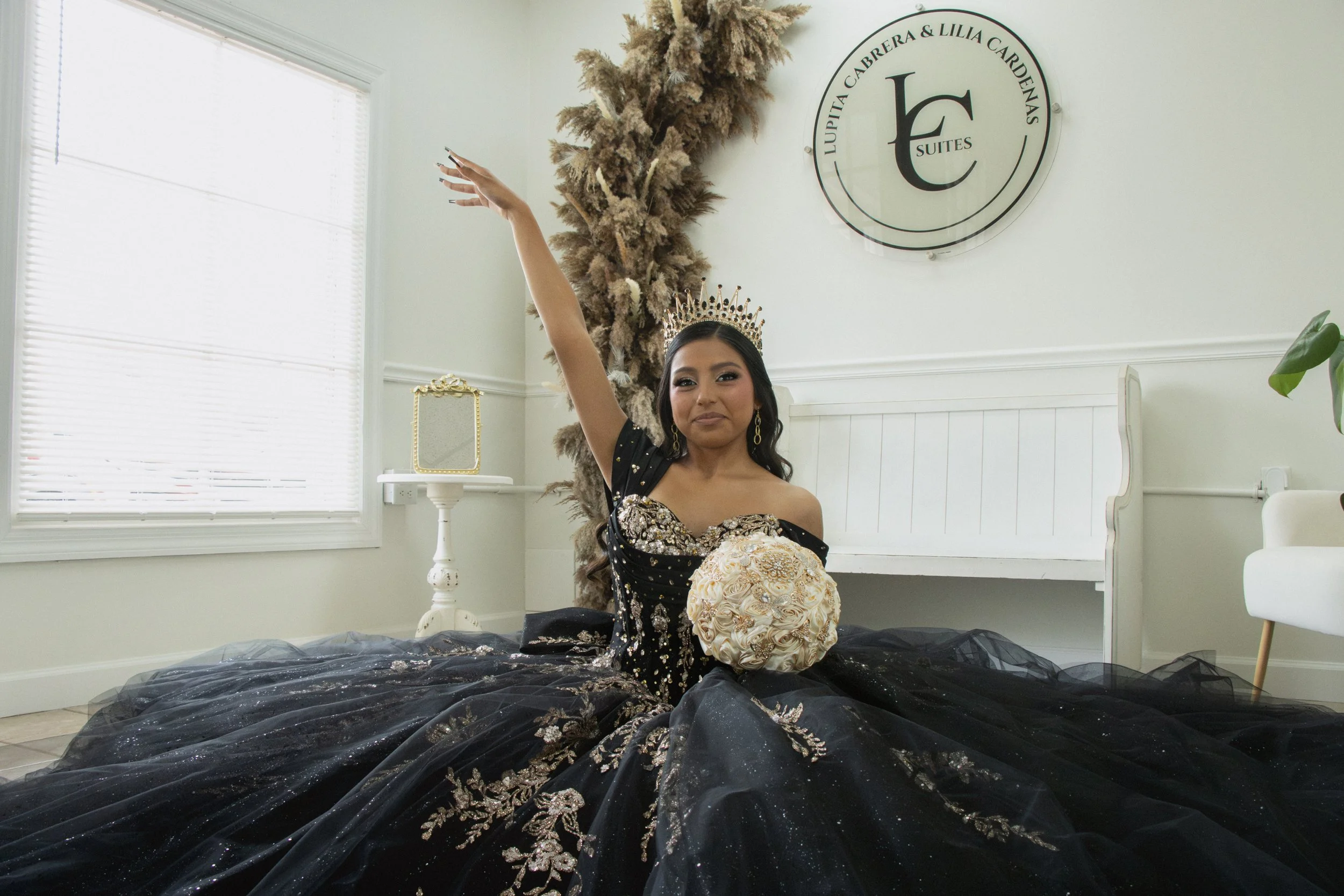 A woman wearing a crown and a black gown with floral embellishments is sitting on the floor with a large, black, floral-embellished skirt spread around her. She is holding a bouquet of white roses and has her arm raised. The room has white walls, a w