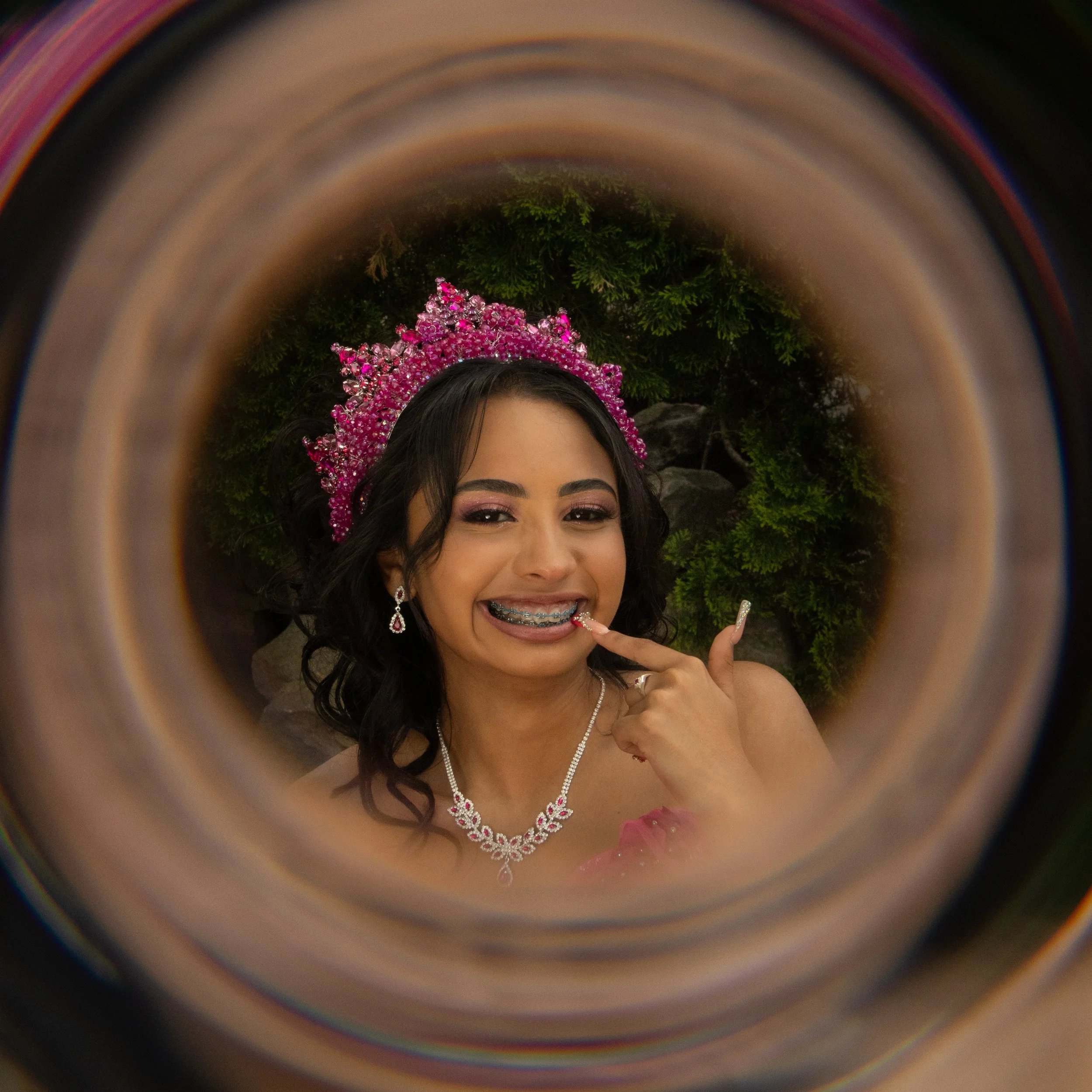 A young woman with dark hair, wearing a pink jeweled crown and jewelry, smiling and showing braces, making a peace sign with her hand