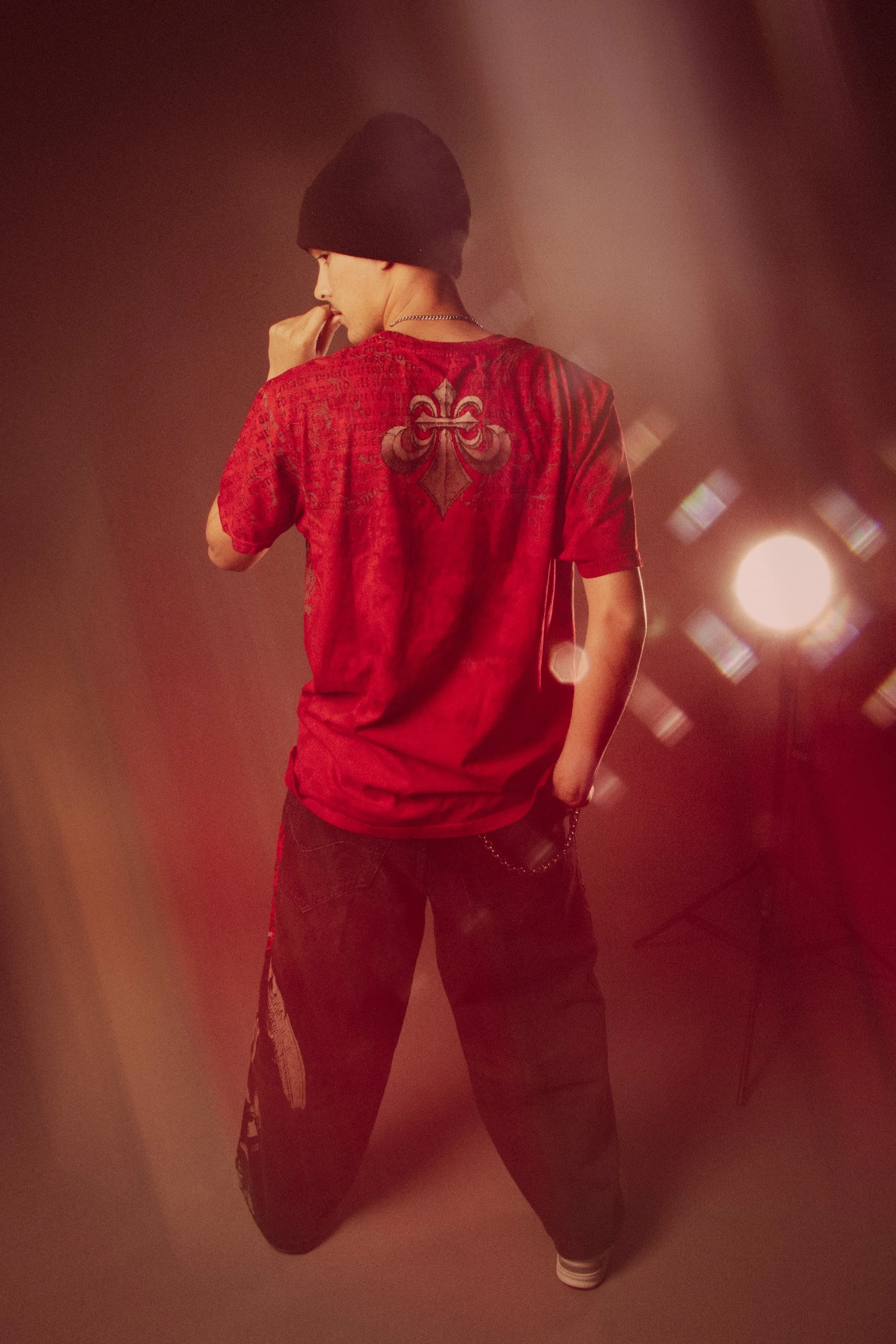 A person wearing a black beanie, red patterned shirt, and black pants standing with one hand near their face, back turned to the camera, with stage or studio lights creating a bright, warm glow.