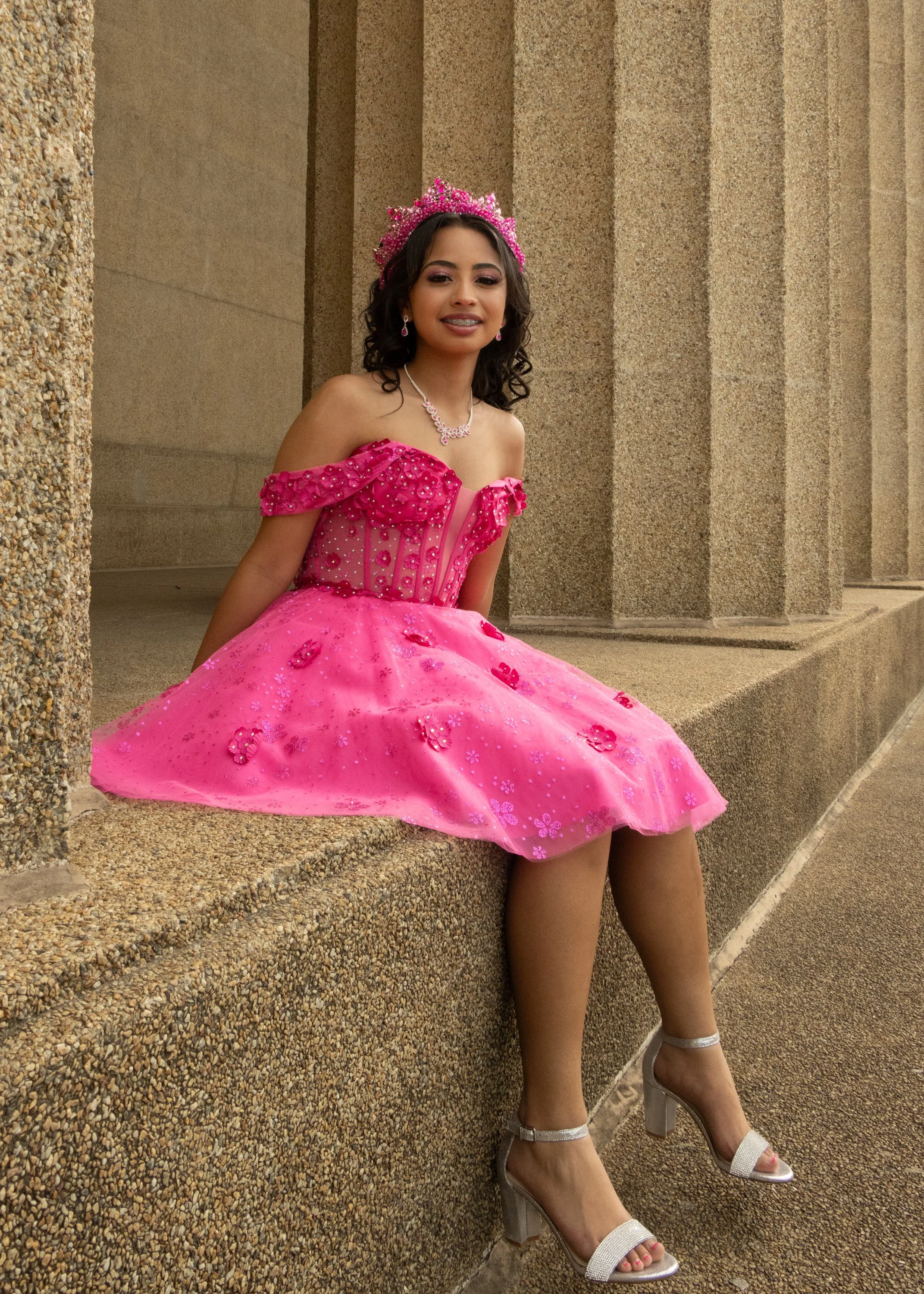 A young woman wearing a pink princess dress with lace and floral details, a pink tiara, jewelry, and silver high heel shoes, sitting on a stone ledge against a textured stone wall.