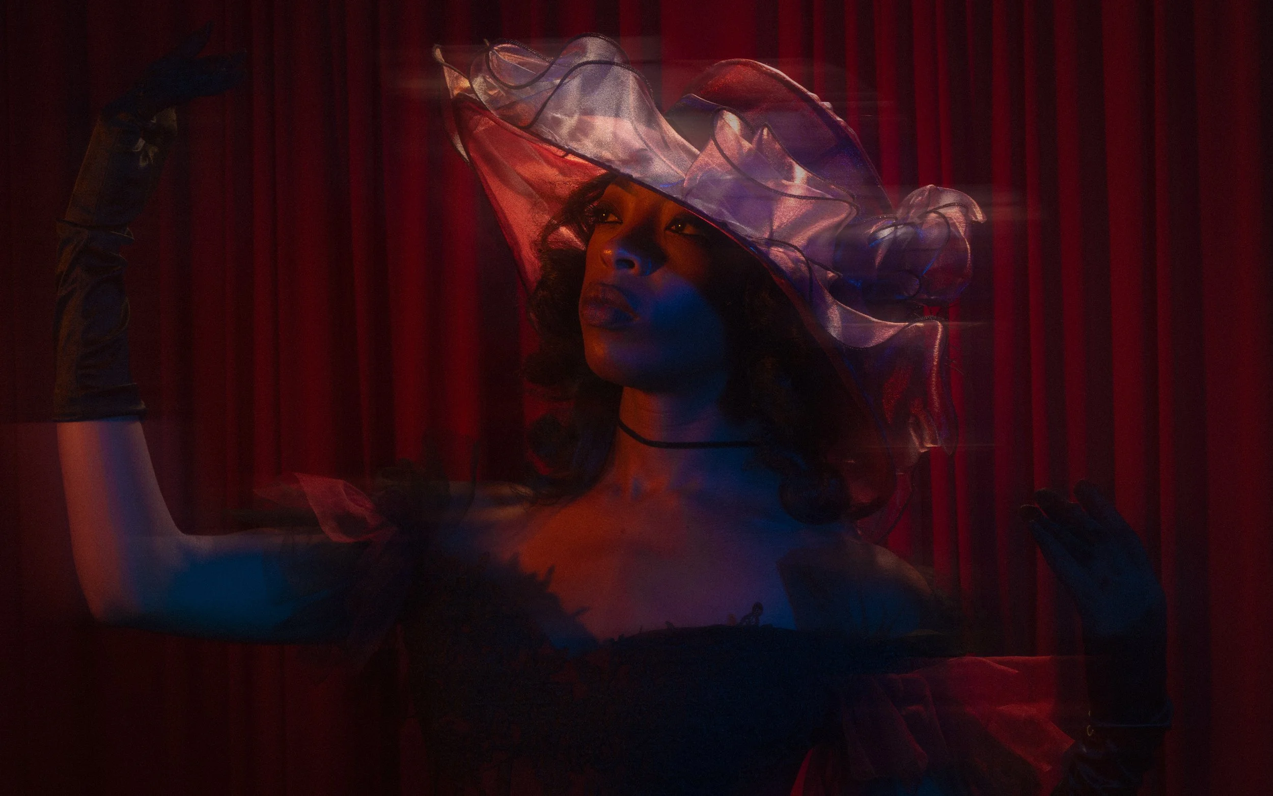A woman wearing a large, transparent, ruffled hat with red accents, illuminated by red and blue lighting, with a red curtain backdrop.