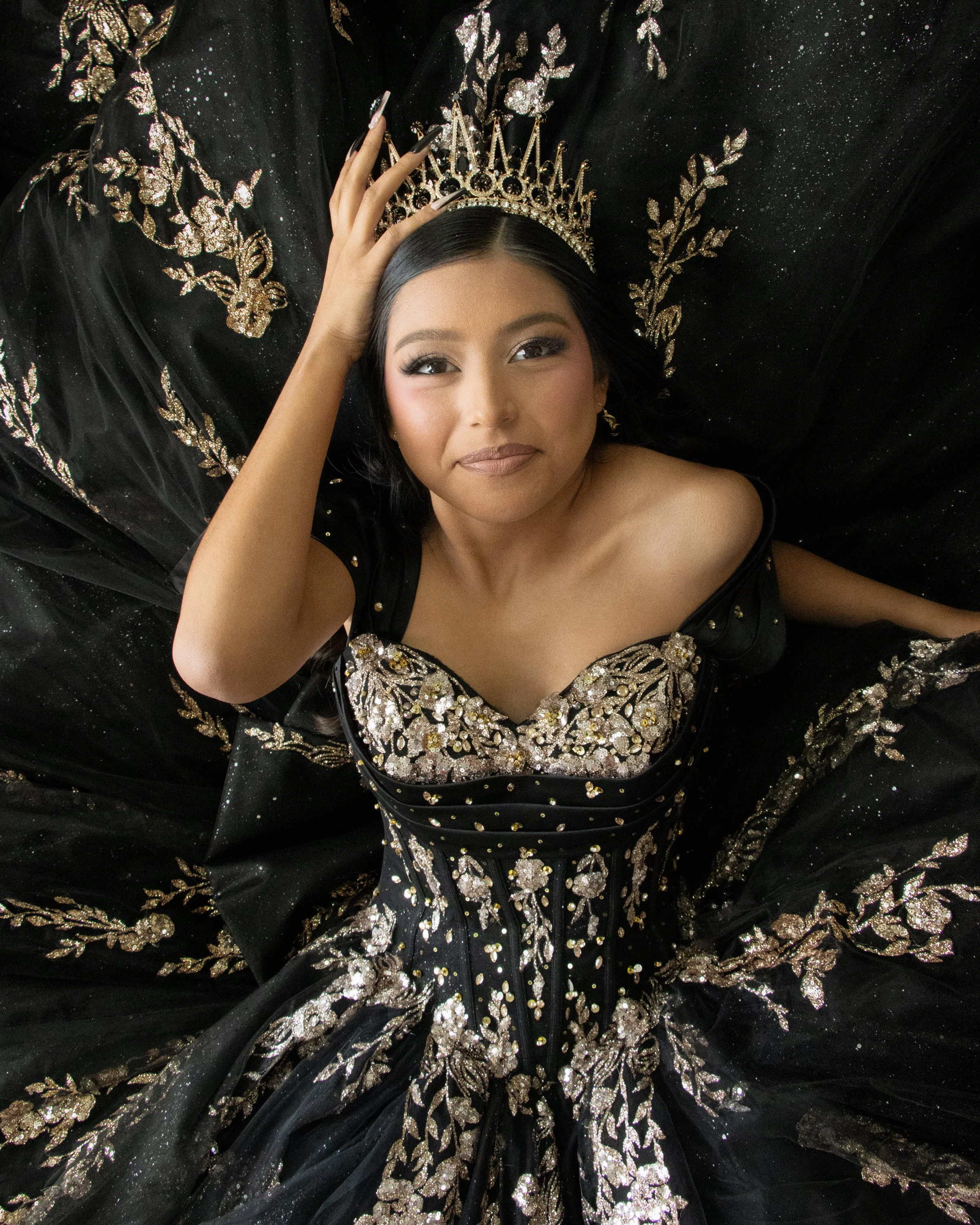 A woman lying on a black, glittery dress with gold embroidery and a gold jeweled crown on her head, looking upwards with a serene expression.