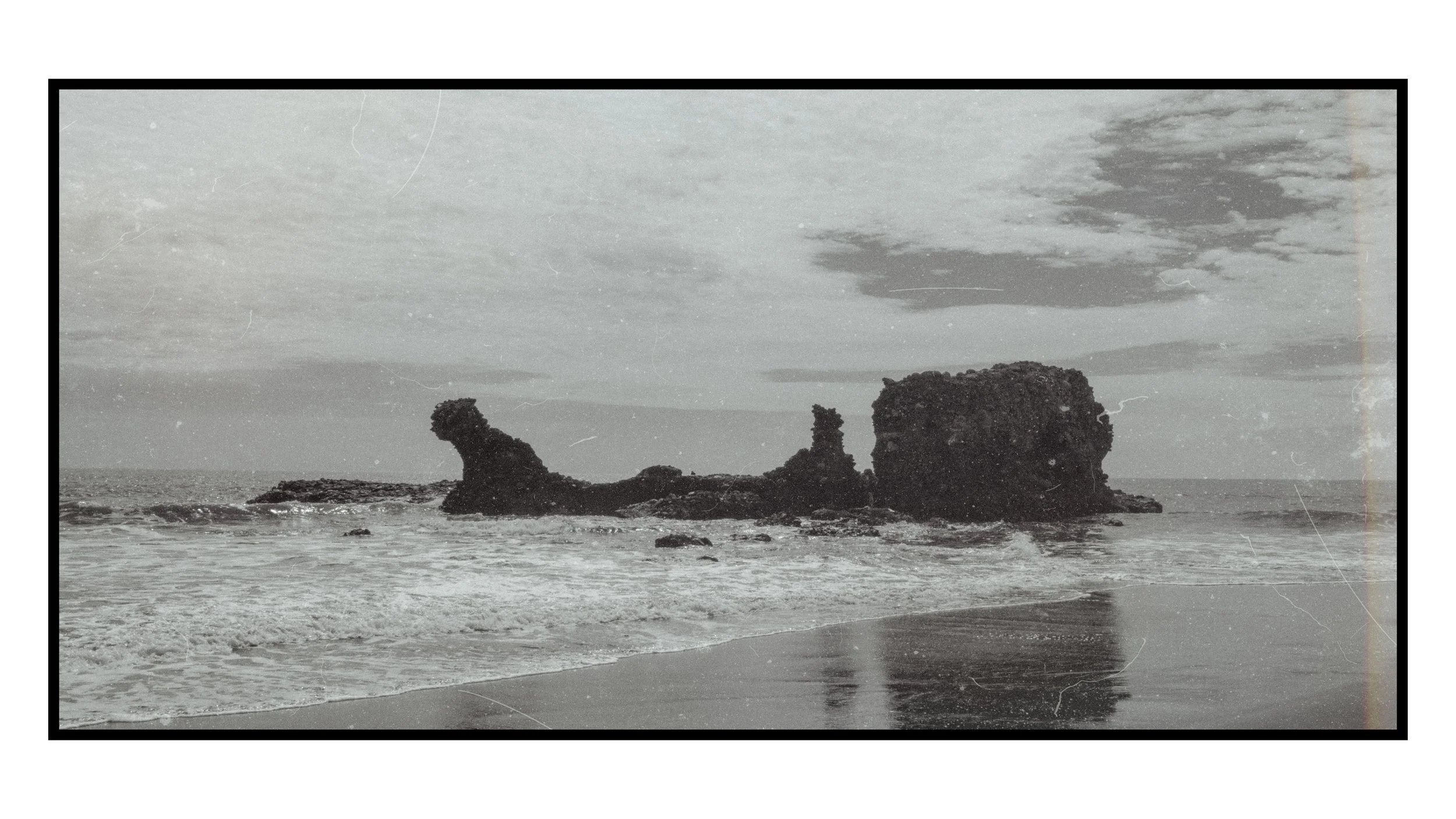 Black and white photo of a rocky coastline with large rock formations in the water and a cloudy sky.