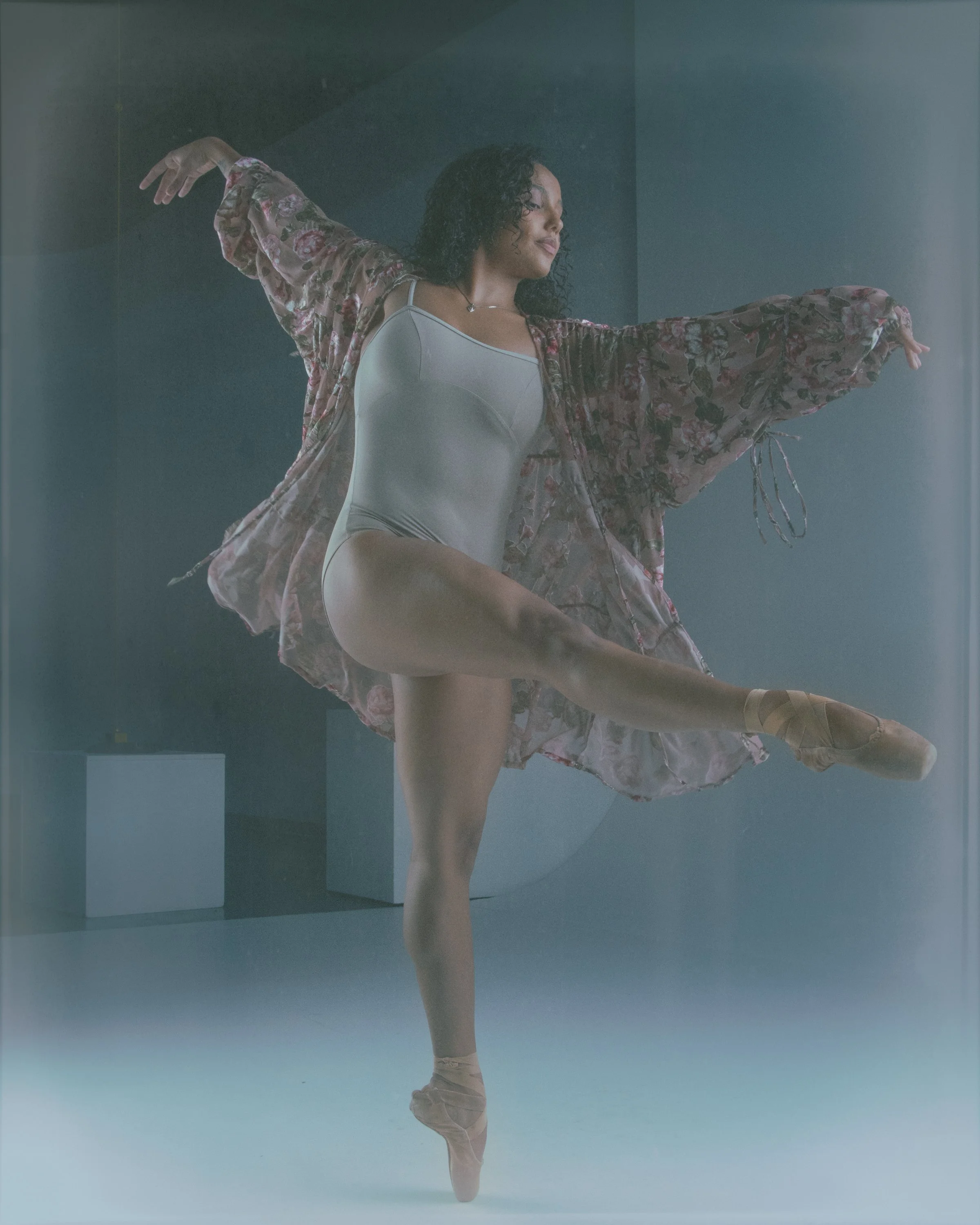 A ballerina in a pose with one leg extended, wearing pointe shoes, a leotard, and a floral kimono-style wrap, against a gray background.
