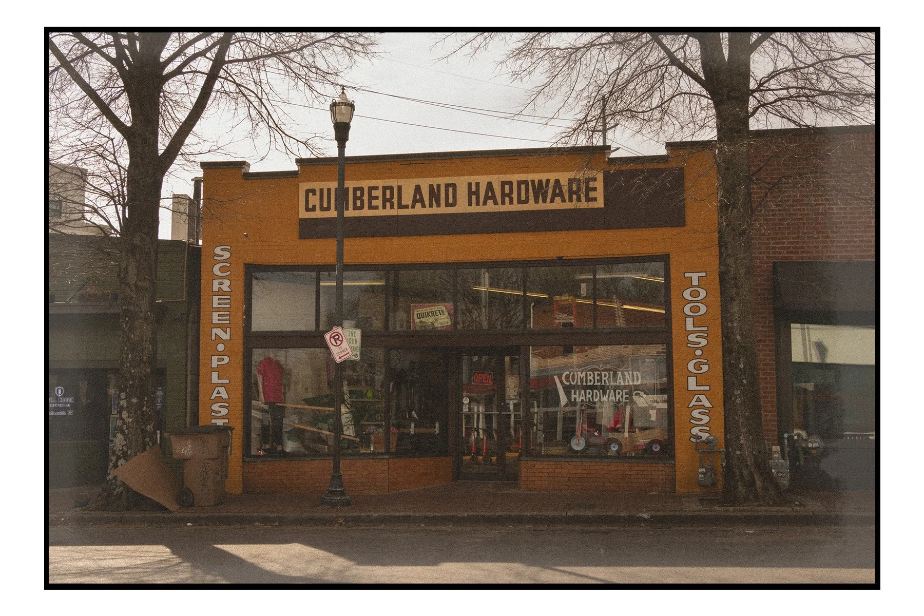 Front view of Cumberland Hardware store with large yellow and black signage, two leafless trees on either side, and a street lamp in front.