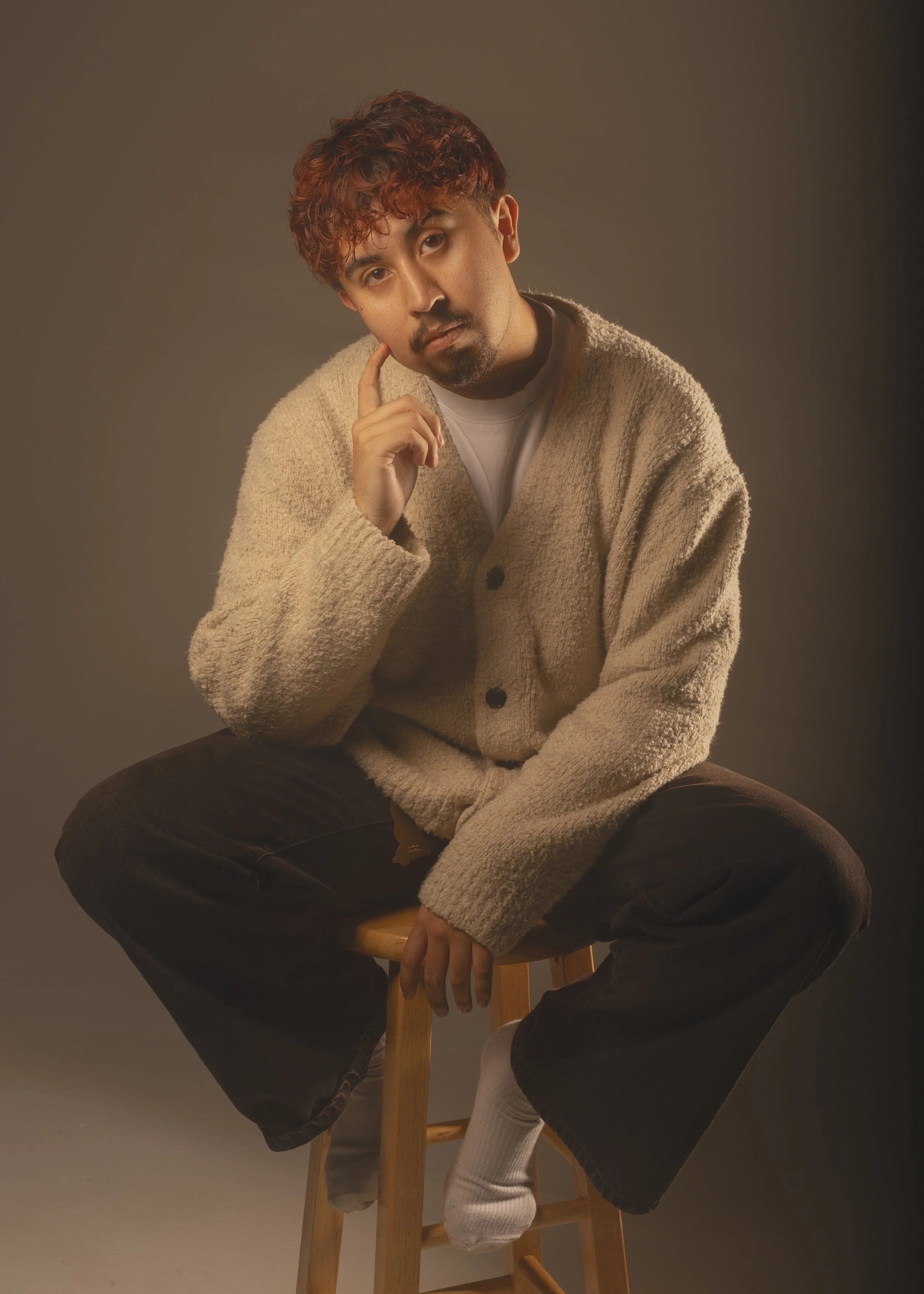 A man with curly reddish hair and a goatee sits on a stool, wearing a beige cardigan, black pants, and white socks. He touches his face with his right hand and looks directly at the camera against a dark, neutral background.