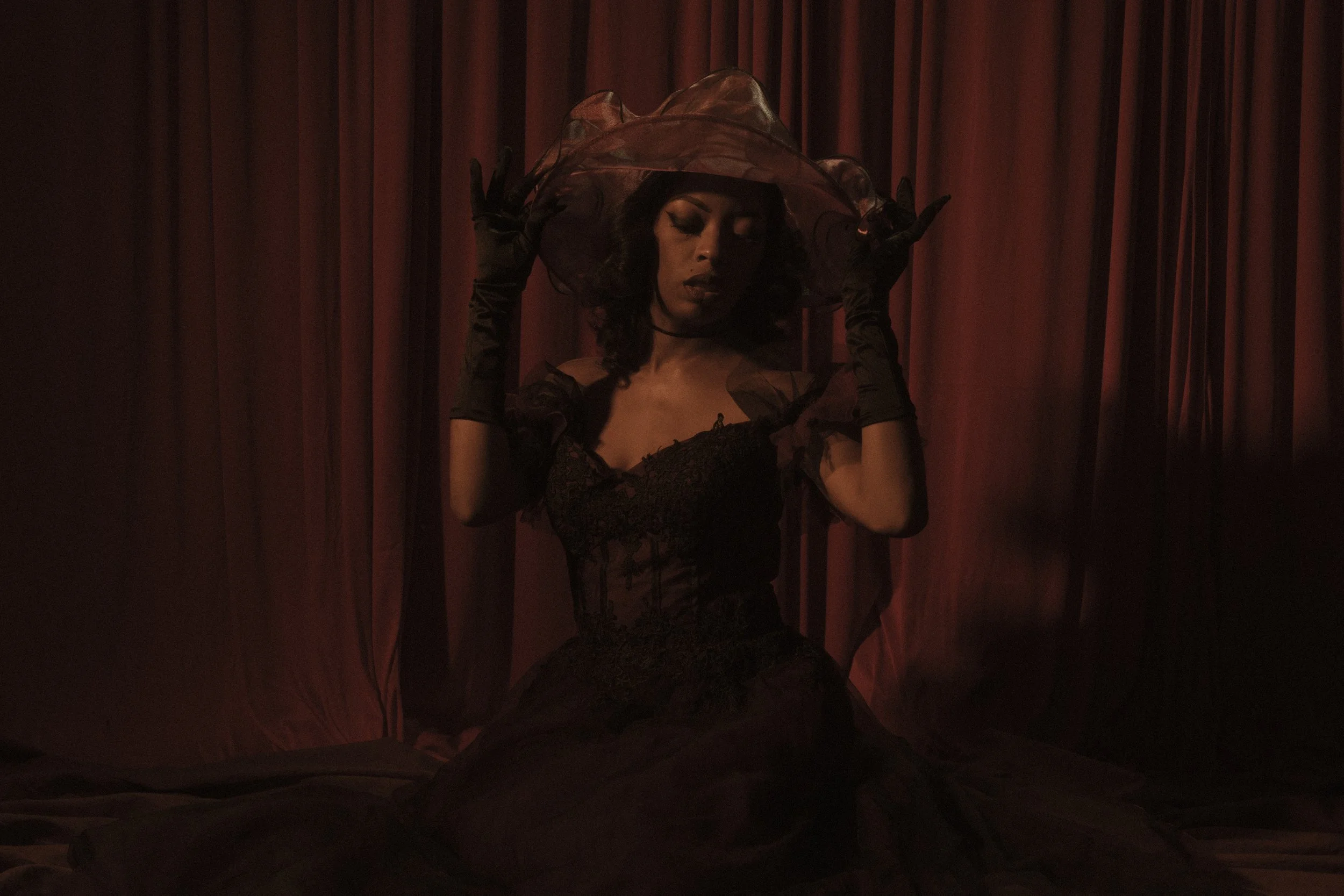 A woman dressed in black with a lace dress, gloves, and a large sheer black hat, standing in front of red curtains, posing with her eyes closed and hands near her head.
