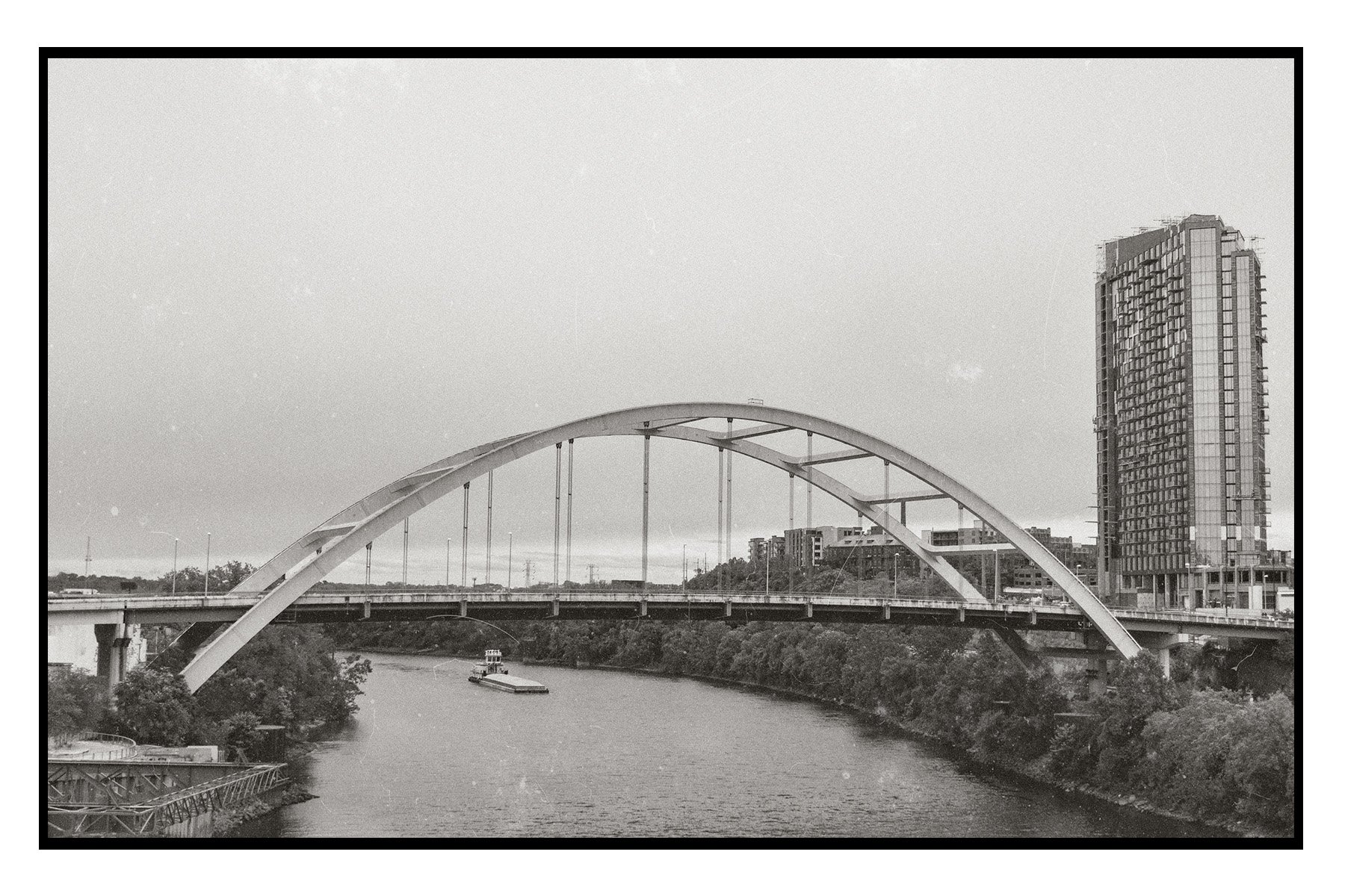 A black-and-white photo of a river with a boat, a bridge, and modern buildings in the background, including a tall, curved skyscraper.