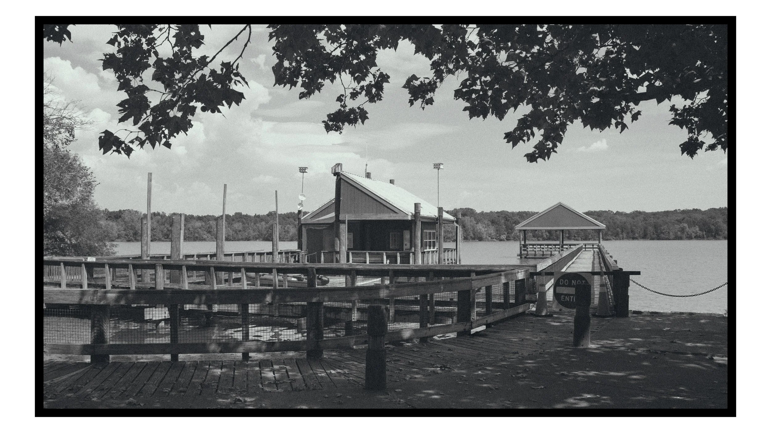 A black-and-white photo of a lakeside dock with small buildings, a pier, and trees overhead.