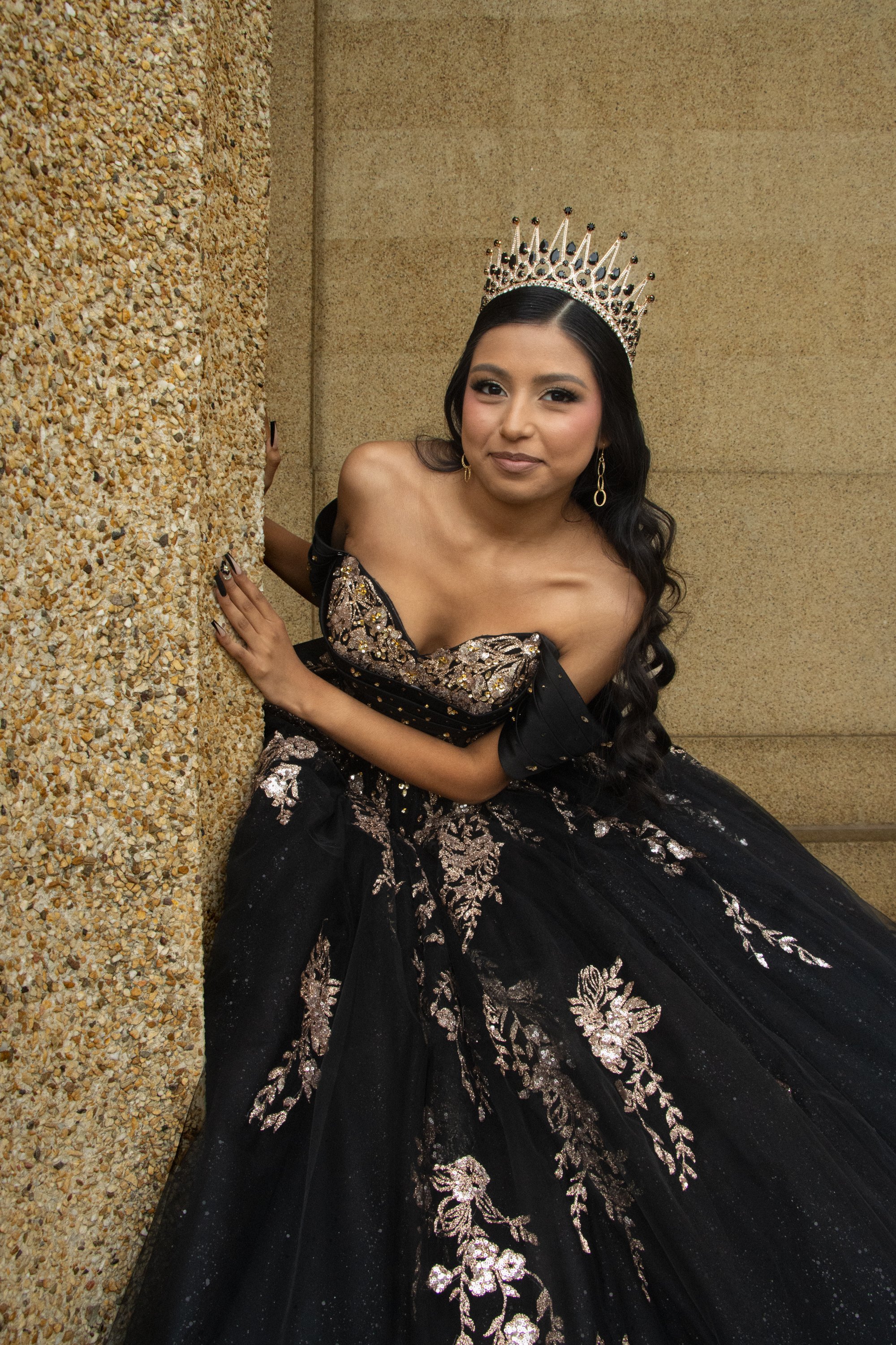 Young woman in a black gown with gold and silver embroidery, wearing a silver crown, sitting on the floor with a speckled textured wall behind her.
