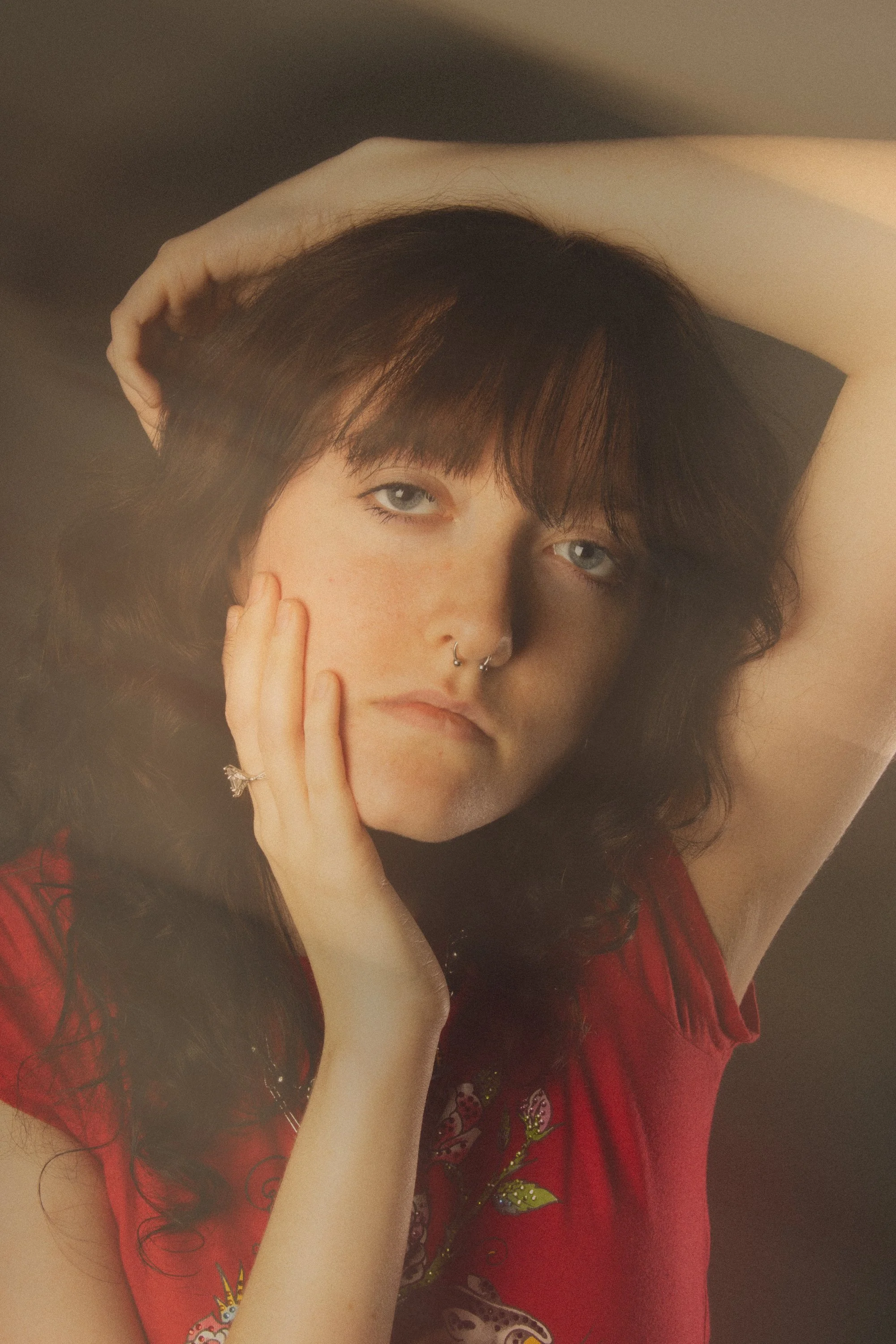 Close-up of a woman with blue eyes and dark brown, wavy hair, wearing a red shirt with embroidered flowers, resting her hand on her face and arm behind her head.