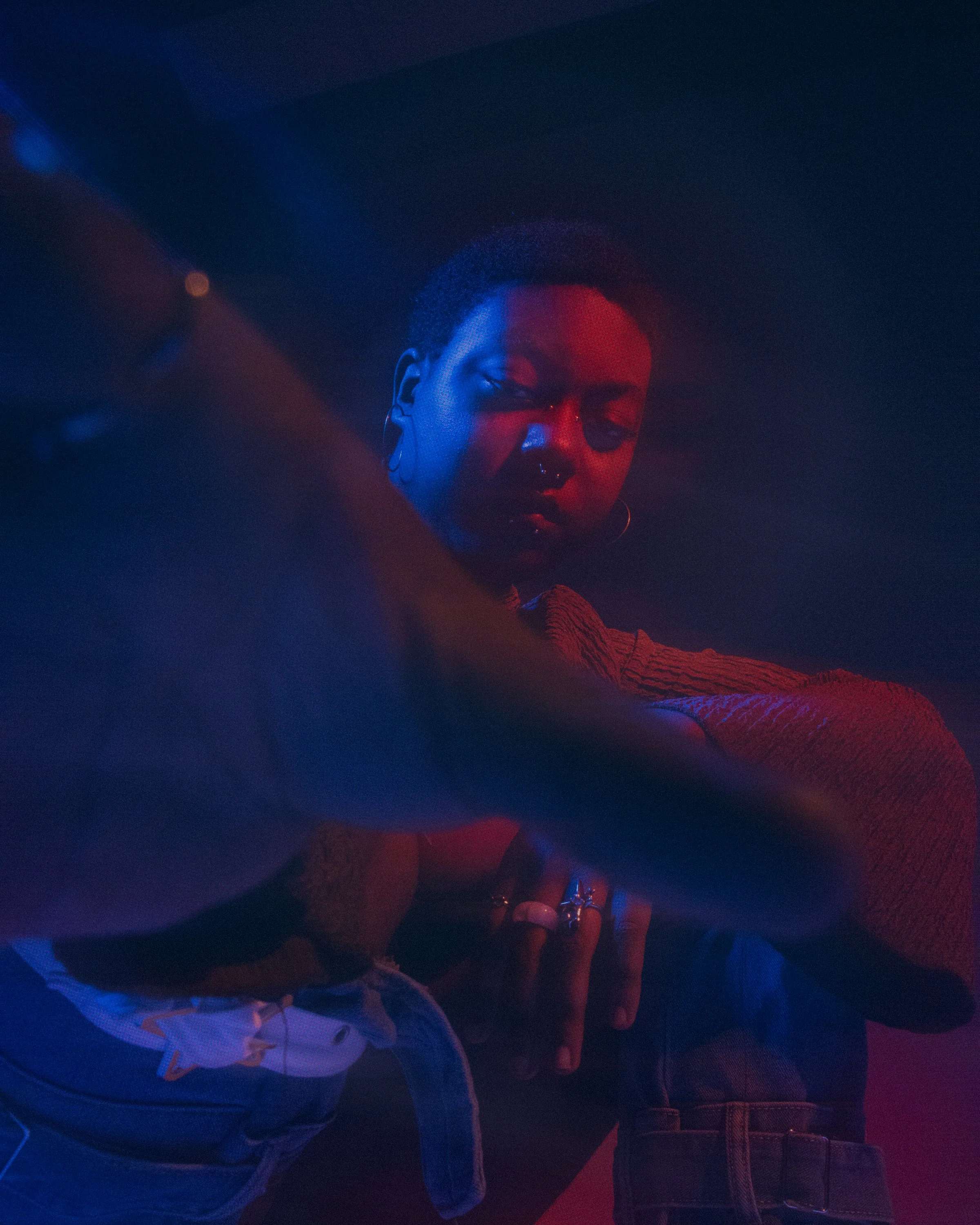 A young person with short curly hair, nose ring, and hoop earrings, appears to be in a dimly lit setting with red and blue lighting. They are wearing a patterned top and denim jacket, with a hand extended into the frame and rings on their fingers.