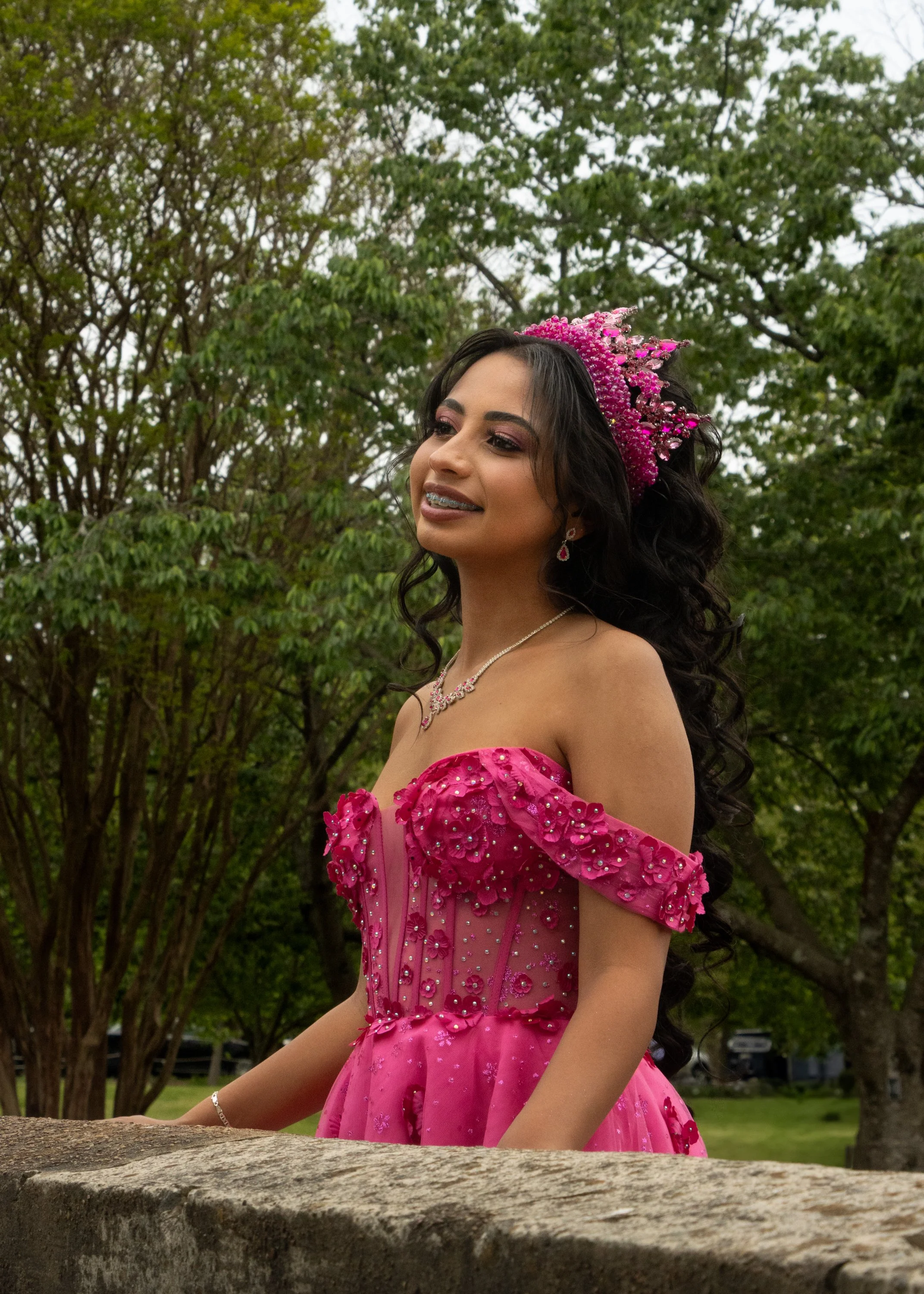 Young woman in a pink, beaded gown and matching tiara standing outdoors in front of green trees.