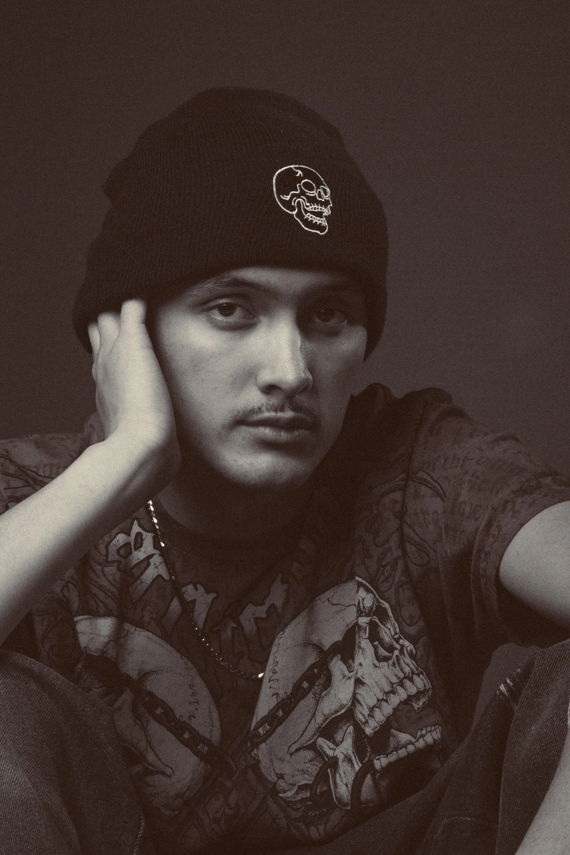 A person wearing a black beanie with a skull logo, a graphic T-shirt with a skull and chains design, and dark jeans, sitting with one hand resting on the head.
