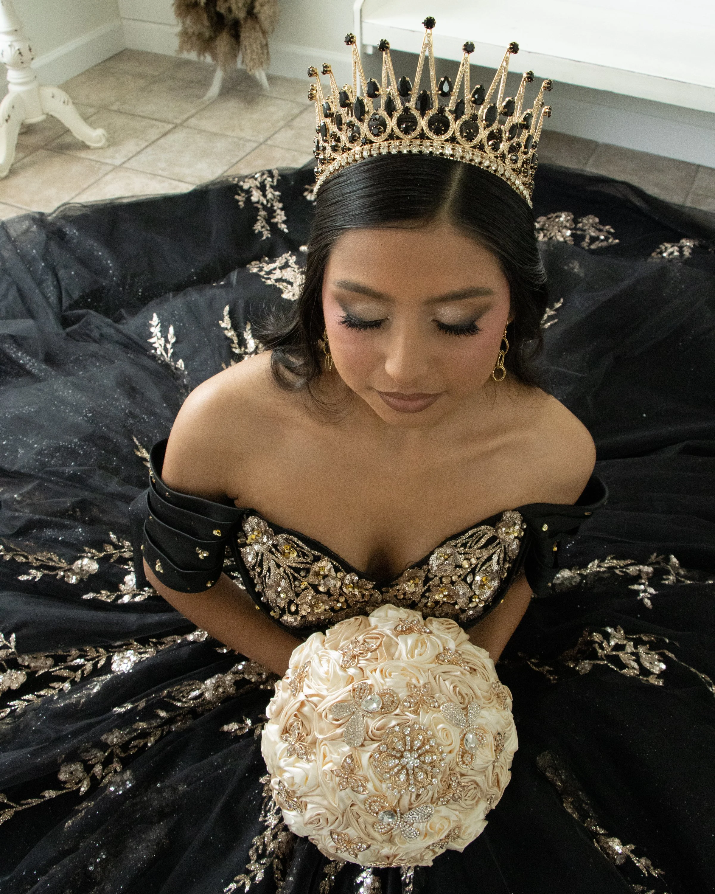 A woman with her eyes closed, wearing a black and gold embellished gown and a crown, sitting on the floor with a bouquet of beige roses in her lap.
