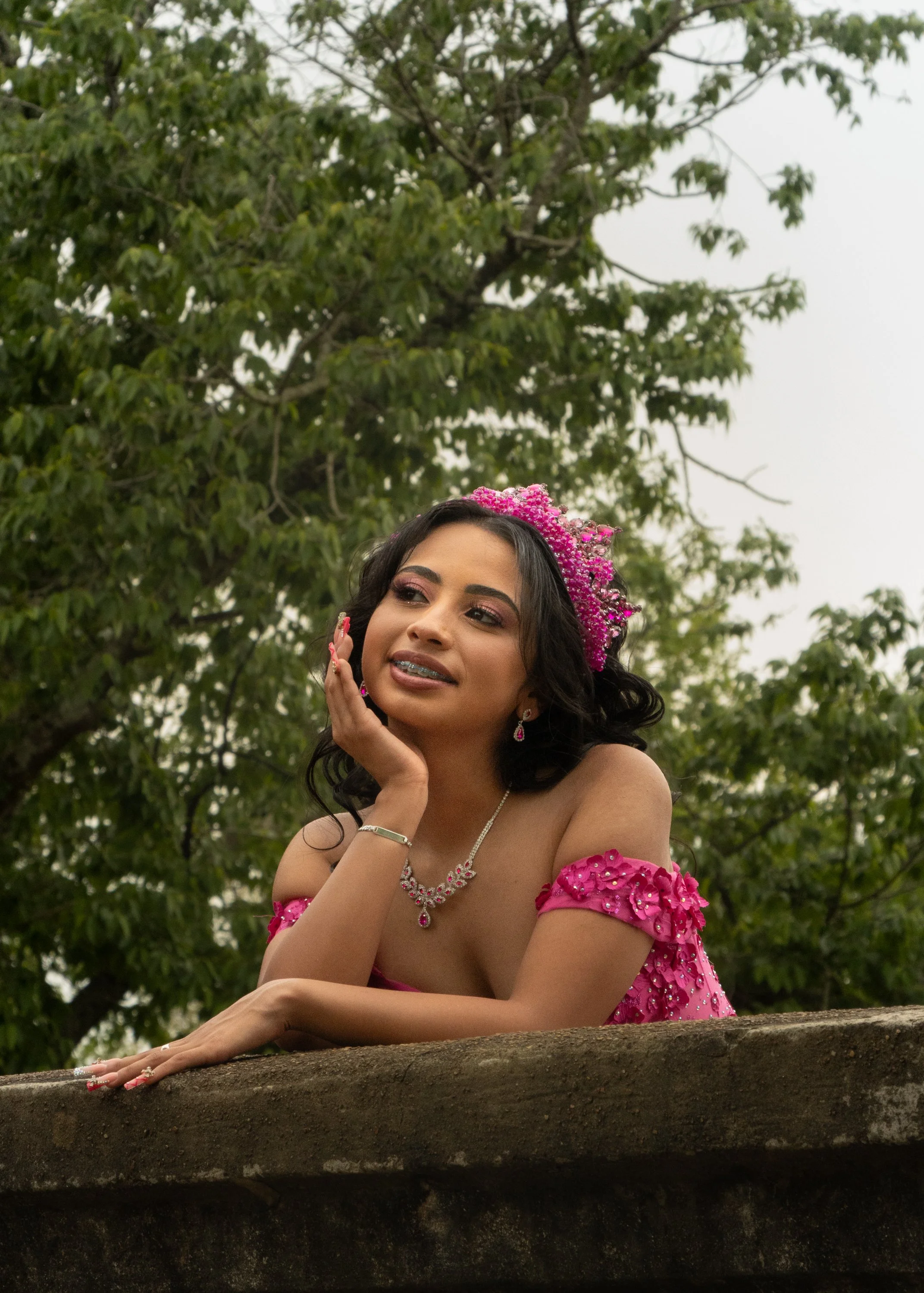 A woman dressed in a pink gown with floral embellishments, wearing matching jewelry and a flower headpiece, rests her hand on her face and looks off into the distance, with lush green trees in the background.