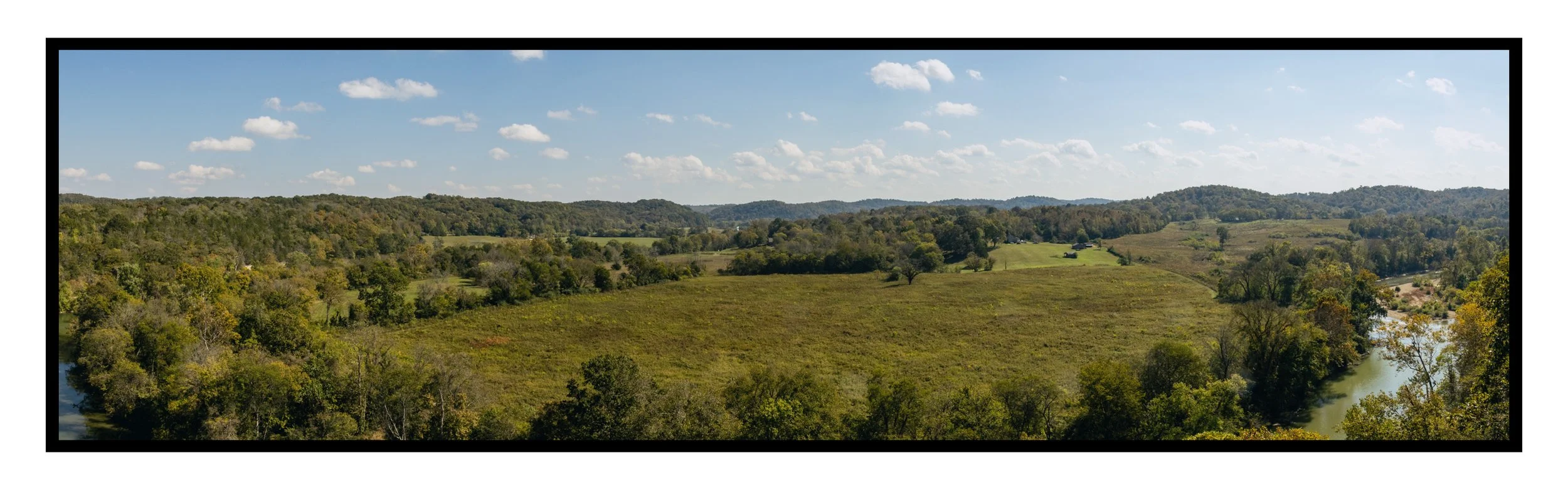 Panoramic view of a rural landscape with rolling hills, scattered trees, and a small river running through the area under a partly cloudy sky.