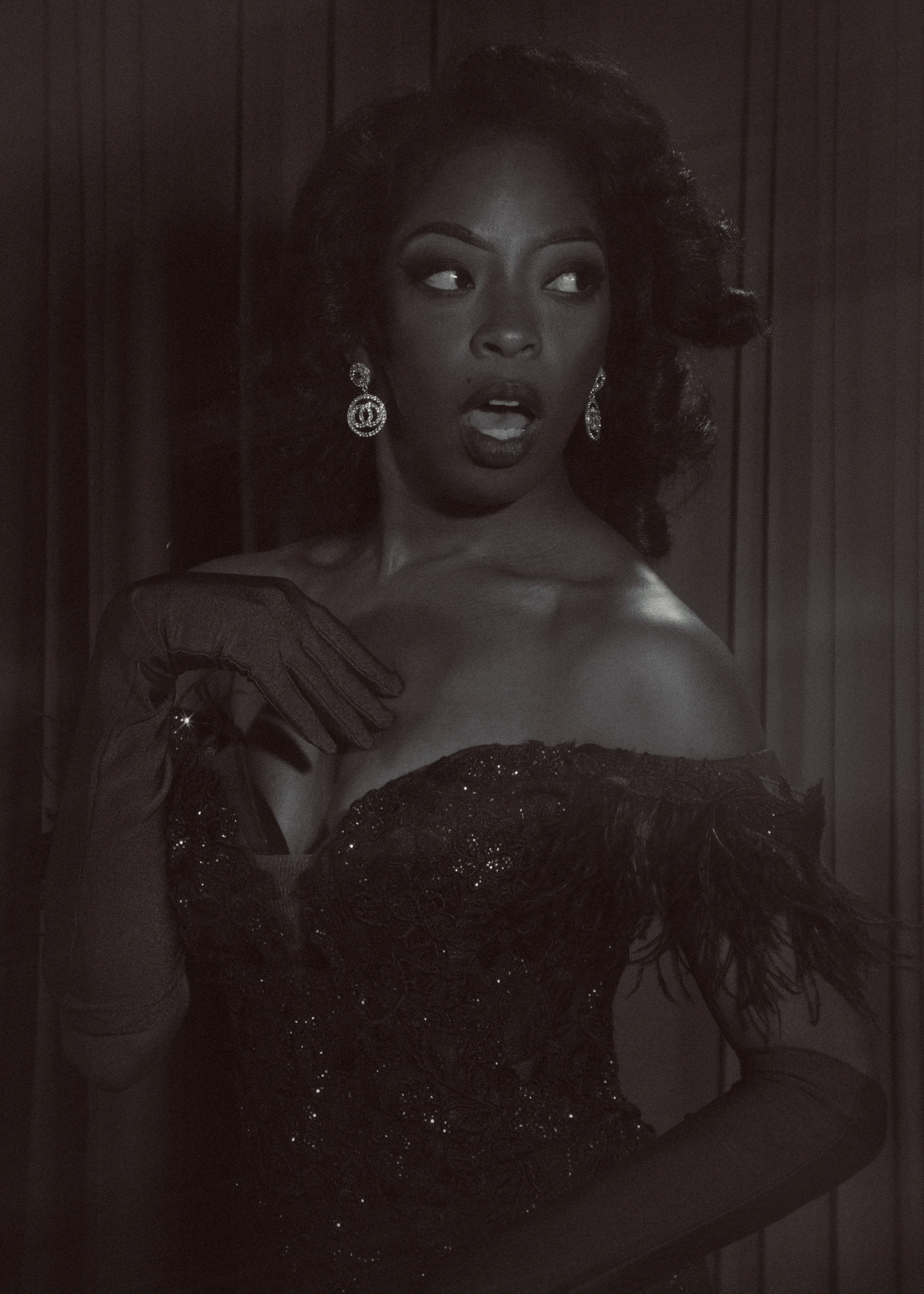Black and white photo of a woman with large earrings, dressed in an elegant off-shoulder dress with lace details, posing against a wooden panel background.