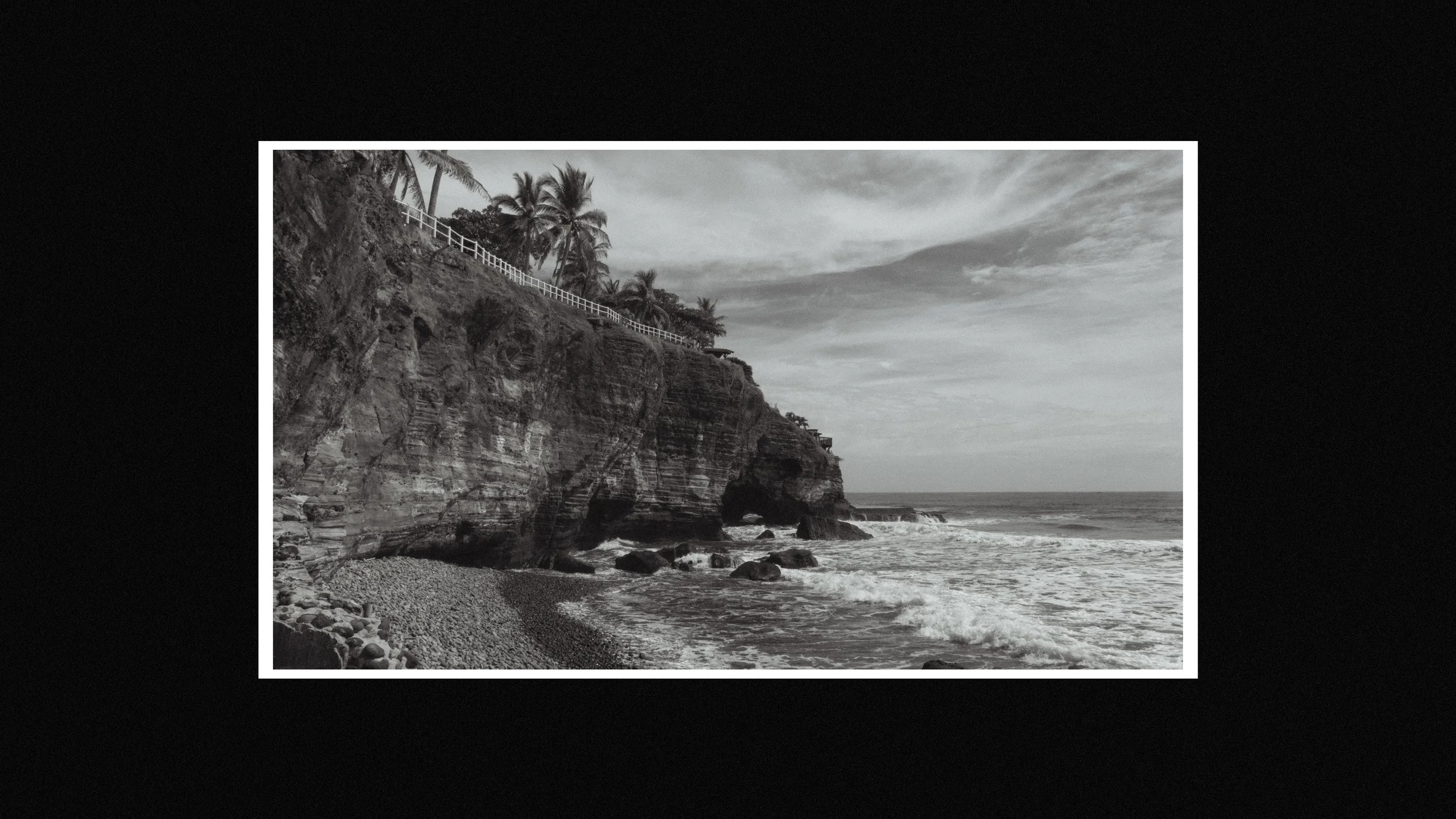 Black and white photo of a rugged cliffside with palm trees and a white fence on top, overlooking the ocean with waves and a rocky beach, under a cloudy sky.