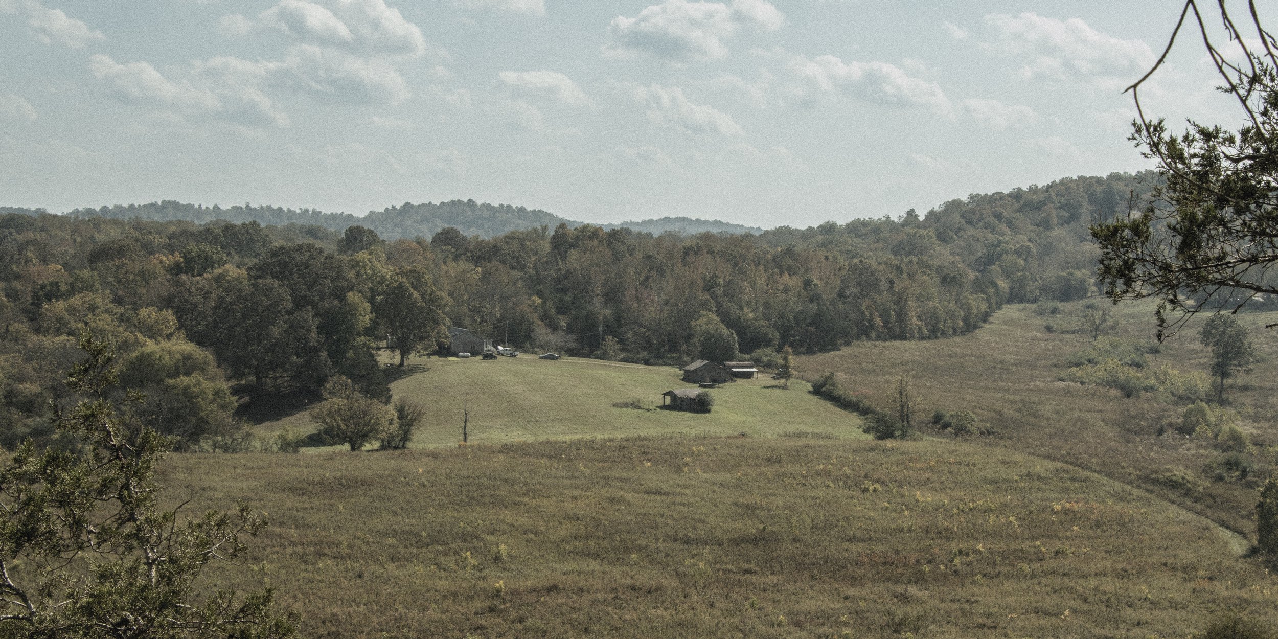 Hilly landscape with trees, grass, and a few buildings on a sunny day.