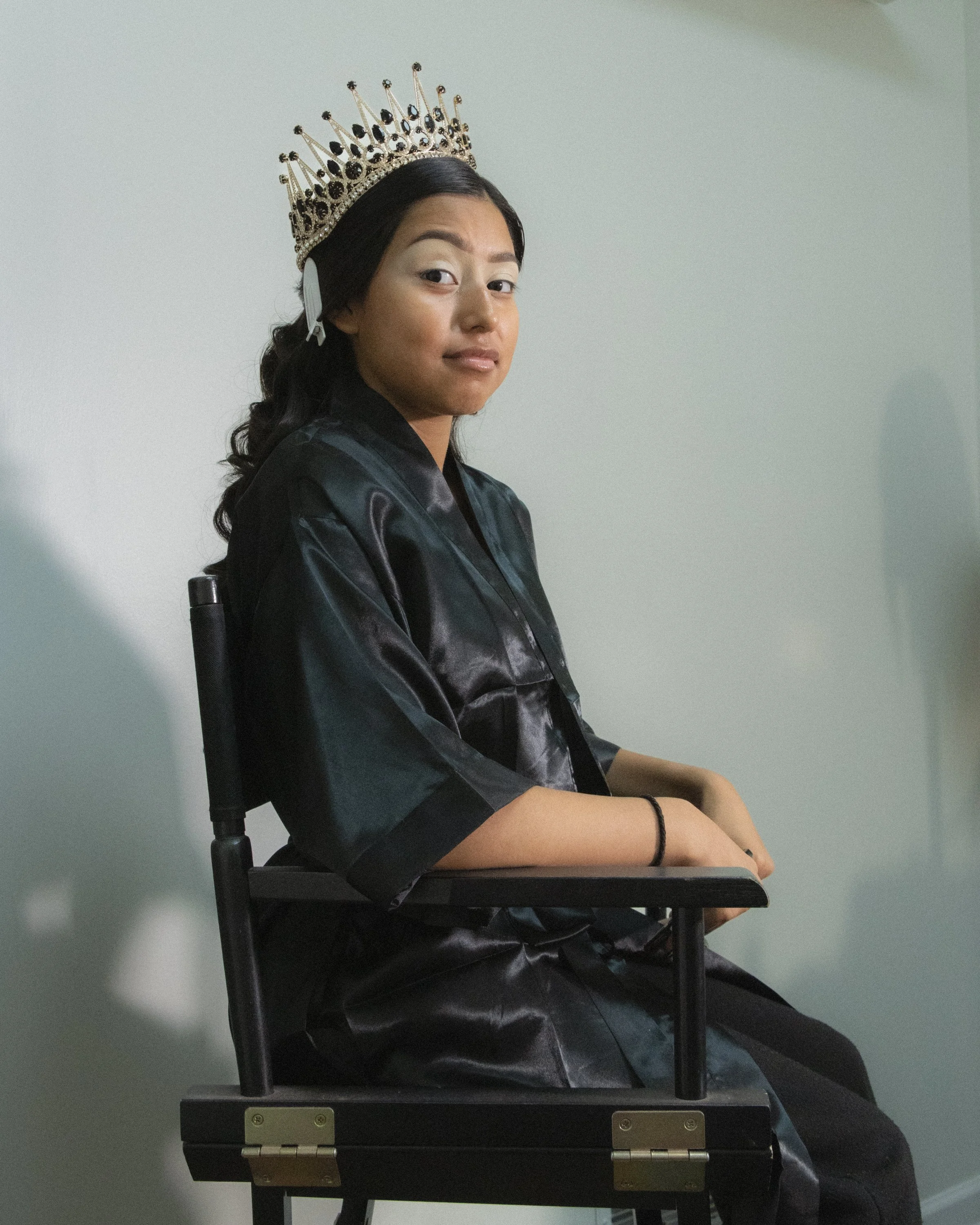 A woman wearing a tiara sitting on a chair, looking at the camera with a slight smile, against a plain white wall background.
