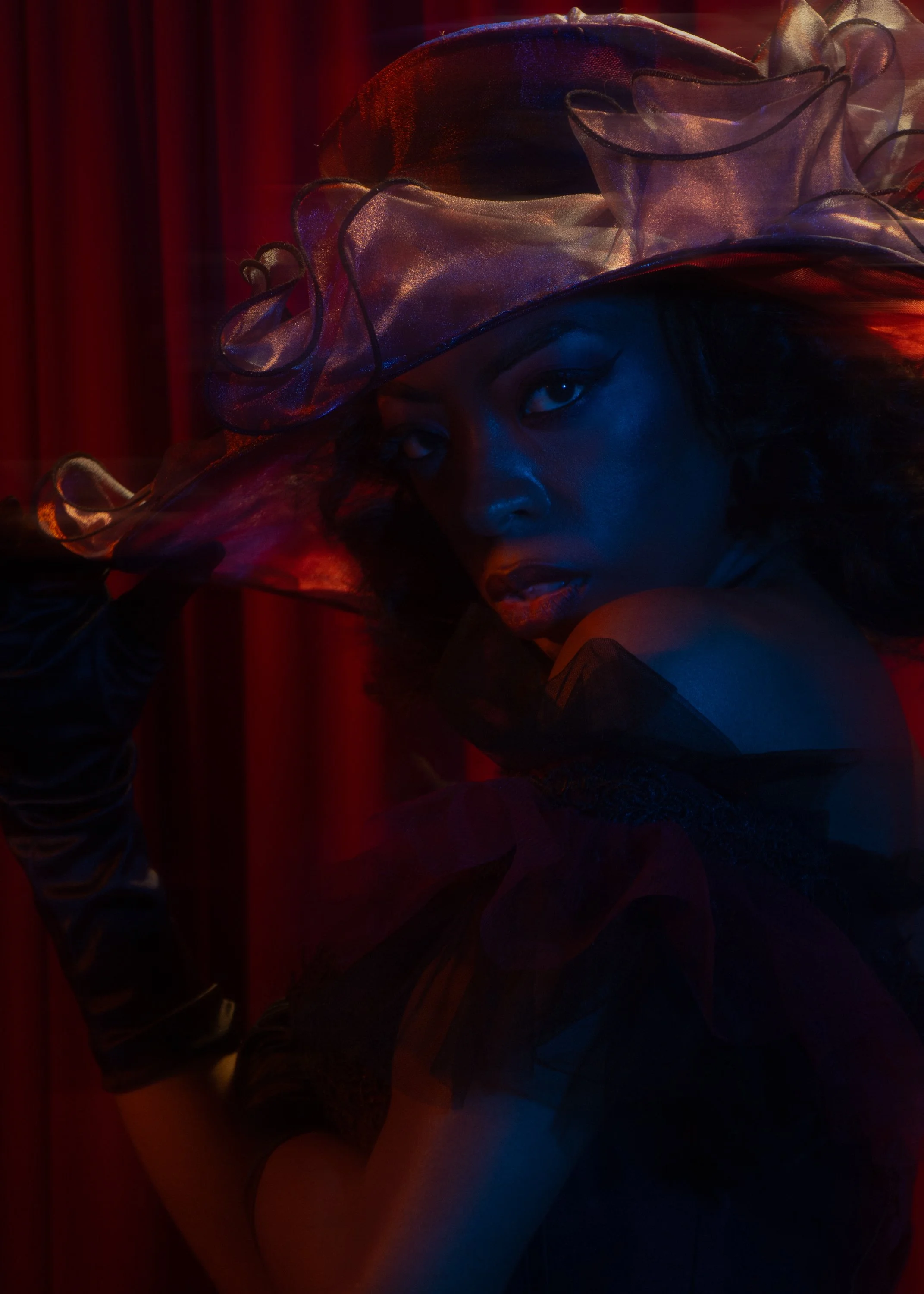 A woman wearing a large, sheer, decorative hat with ribbon accents, looking sideways with dramatic makeup, against a dark red and black background.