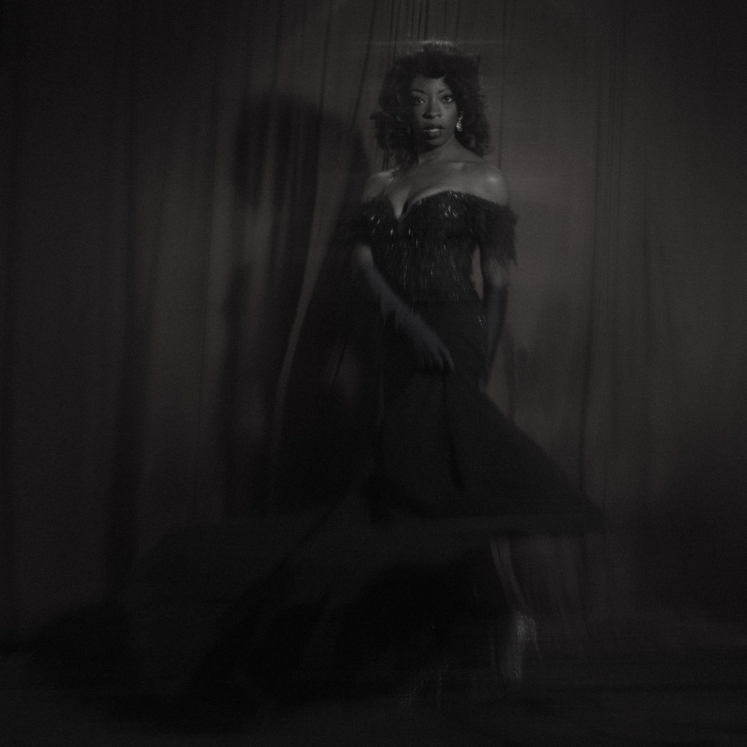 A woman with dark, voluminous hair wearing a strapless black dress with feathered embellishments, standing against a dark background with curtains.