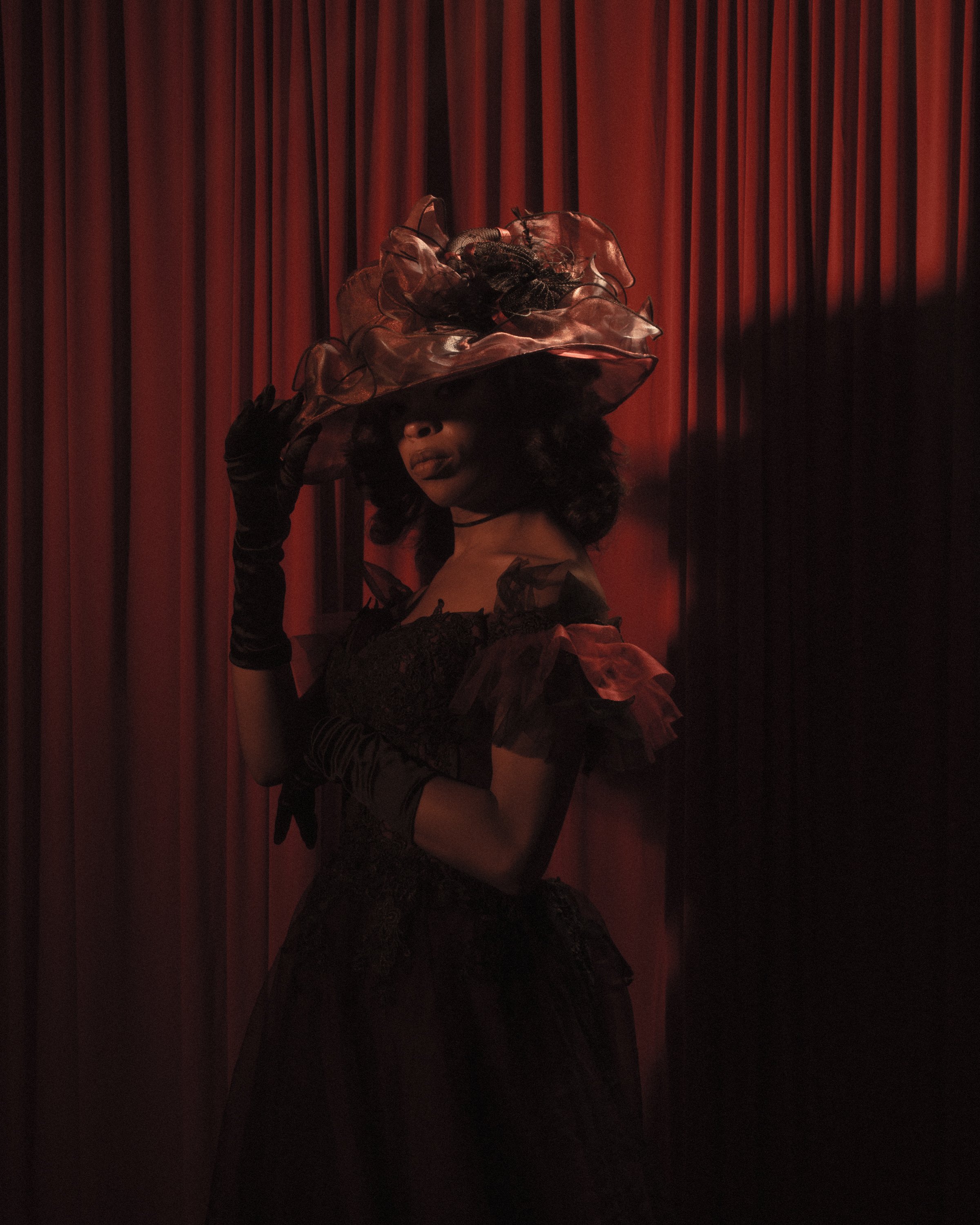 A woman wearing vintage clothing and accessories, including a large ornate hat, standing against a backdrop of red curtains.