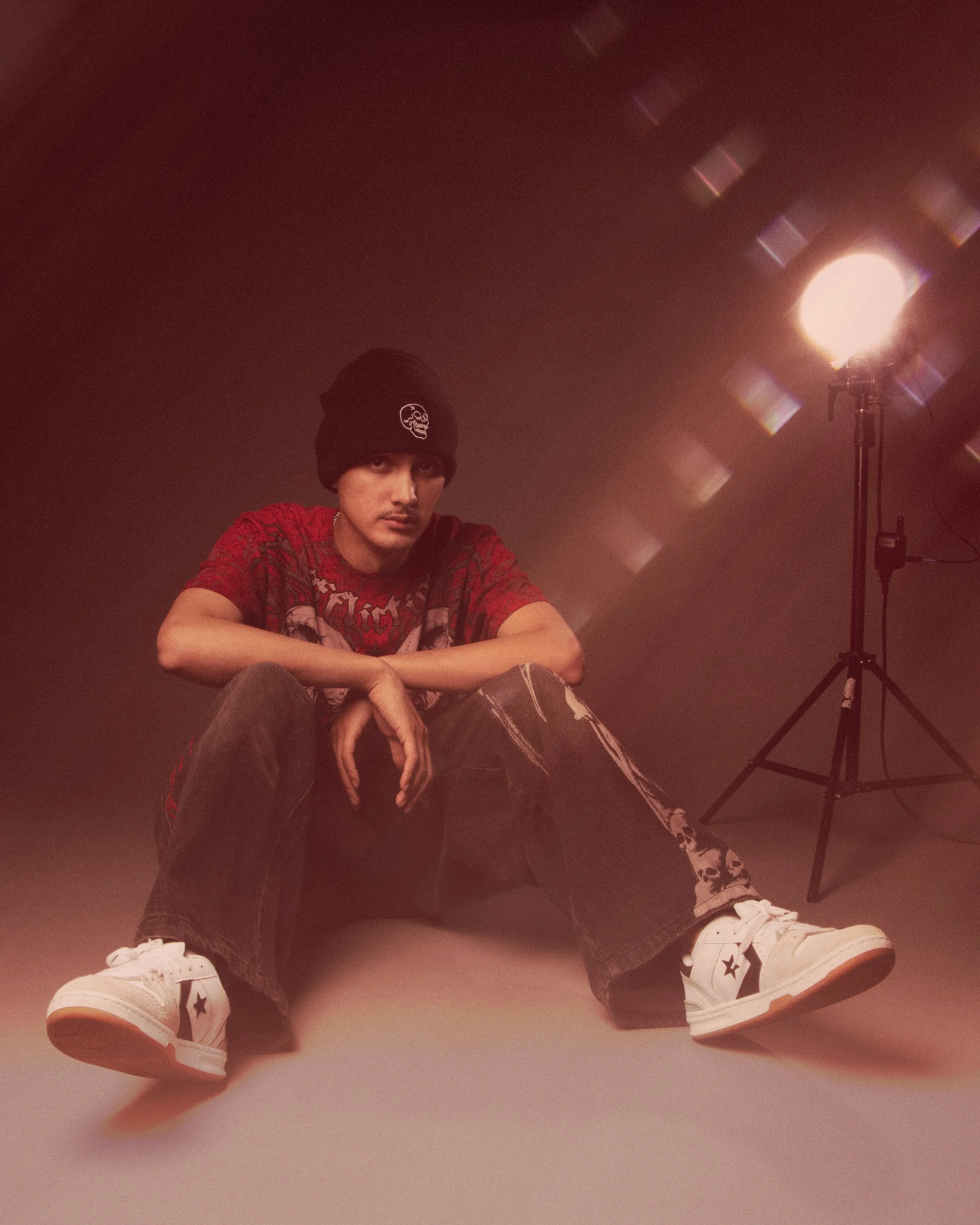 Young man wearing a black beanie, red graphic t-shirt, ripped jeans, and white sneakers, sitting on the floor in front of a light source, with his legs crossed and looking at the camera.
