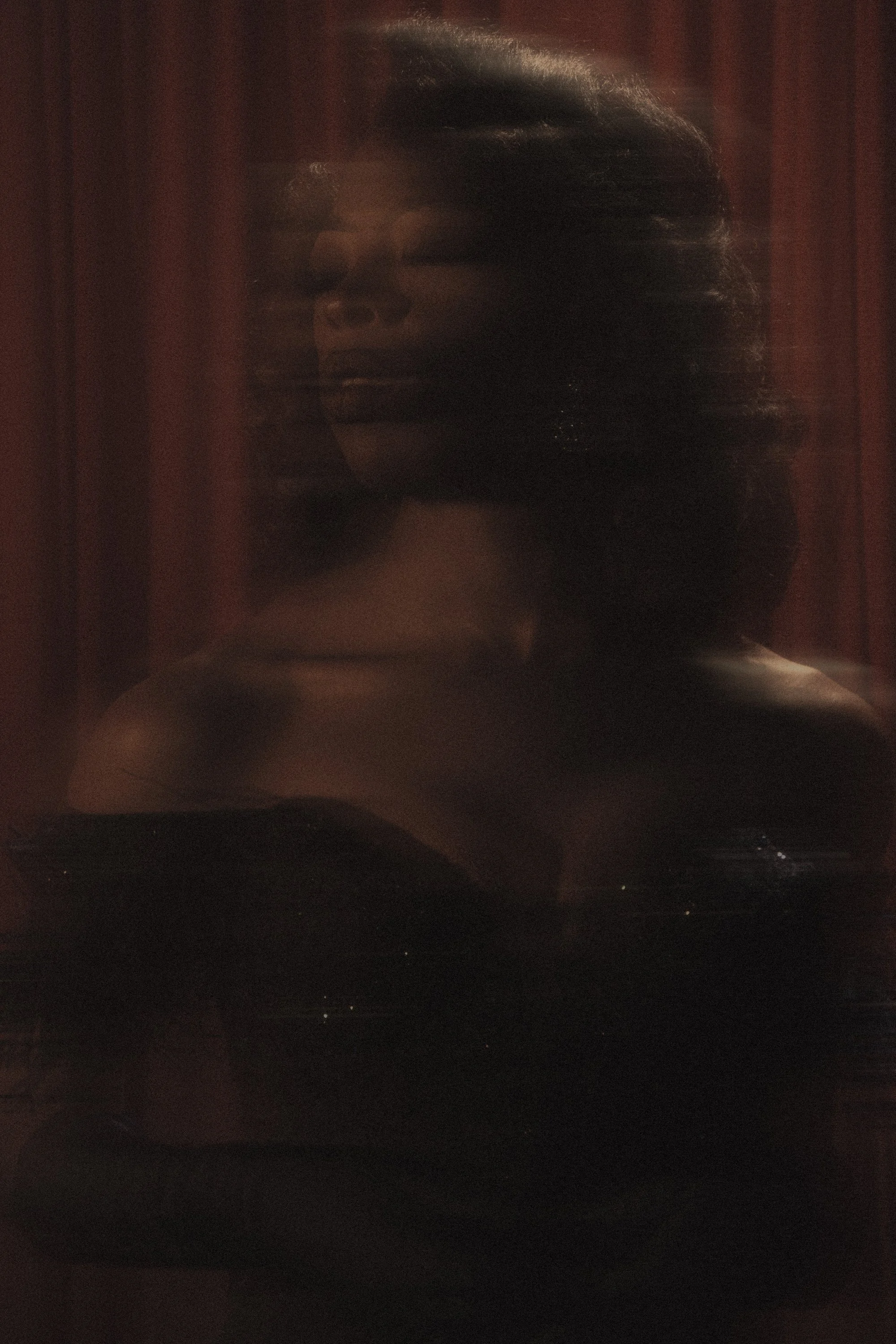 A dark, moody photo of a woman with styled hair and makeup, taken through a reflective or textured surface, with a background of red curtains.