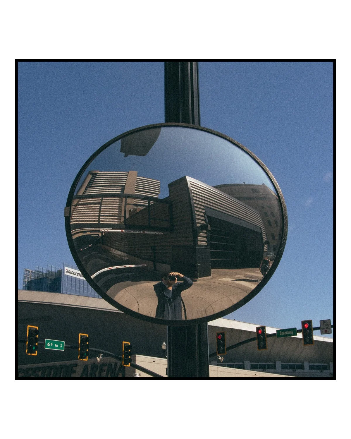 Reflected buildings and sky in a circular mirror mounted on a pole, with a person taking a photo and traffic lights in the foreground.