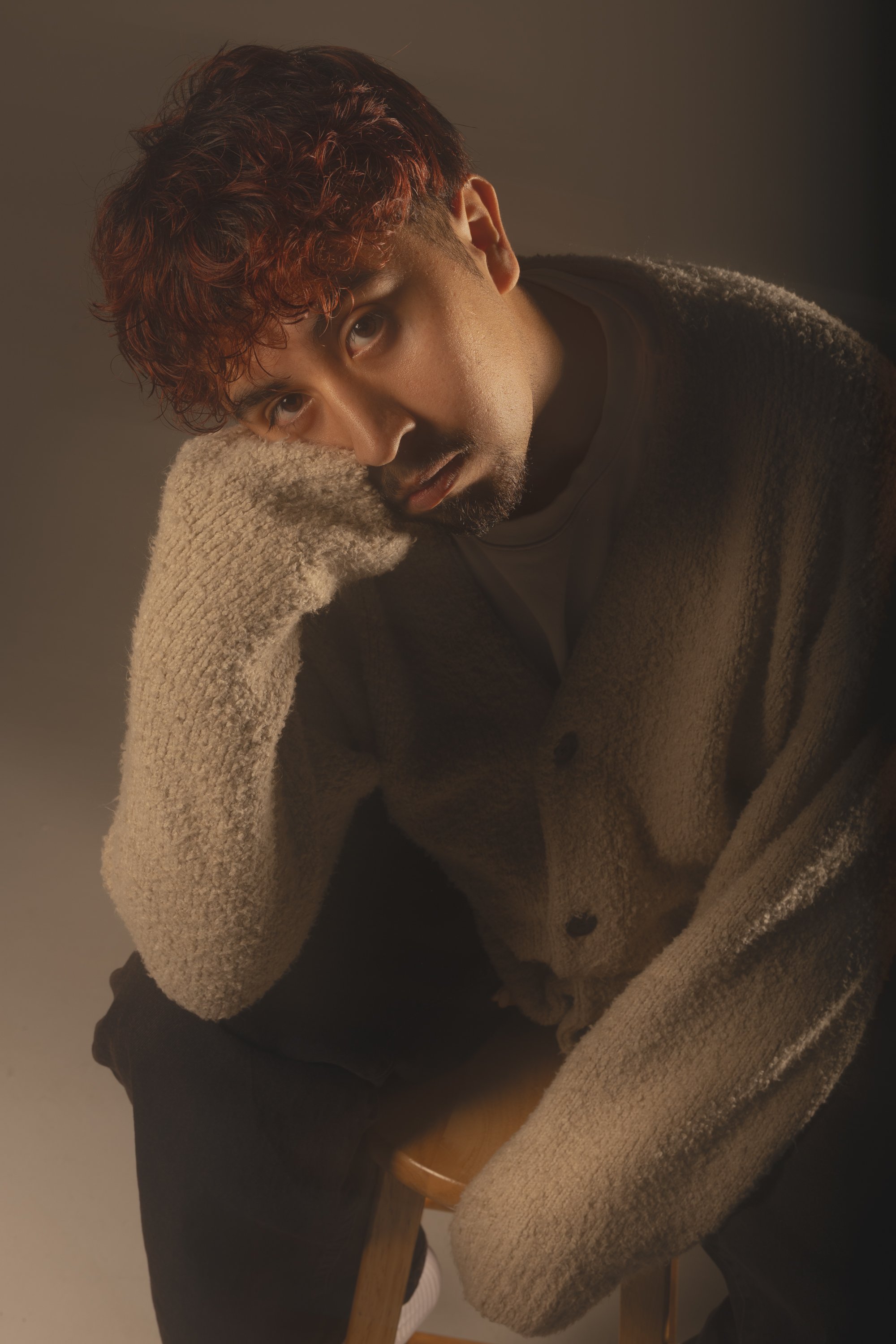 A man with curly hair and a beard, resting his head on his hand. He's wearing a brown coat over a black shirt. The background is plain and gray.