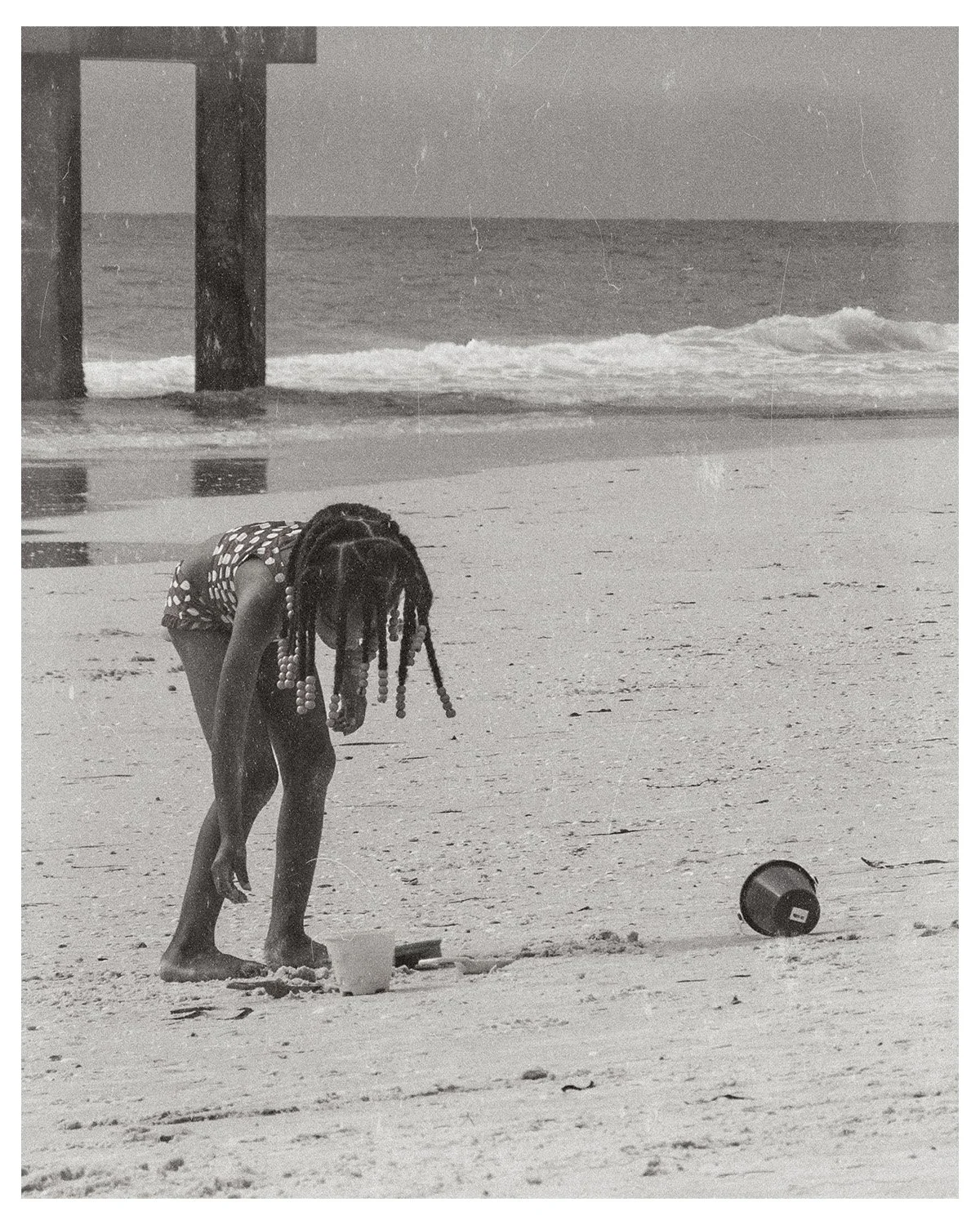 A young girl with braided hair, wearing a polka dot swimsuit, is playing on the beach and bending down to pick something up. There is a bucket and a cup nearby on the sand. The ocean is in the background with waves and a pier visible in the distance.