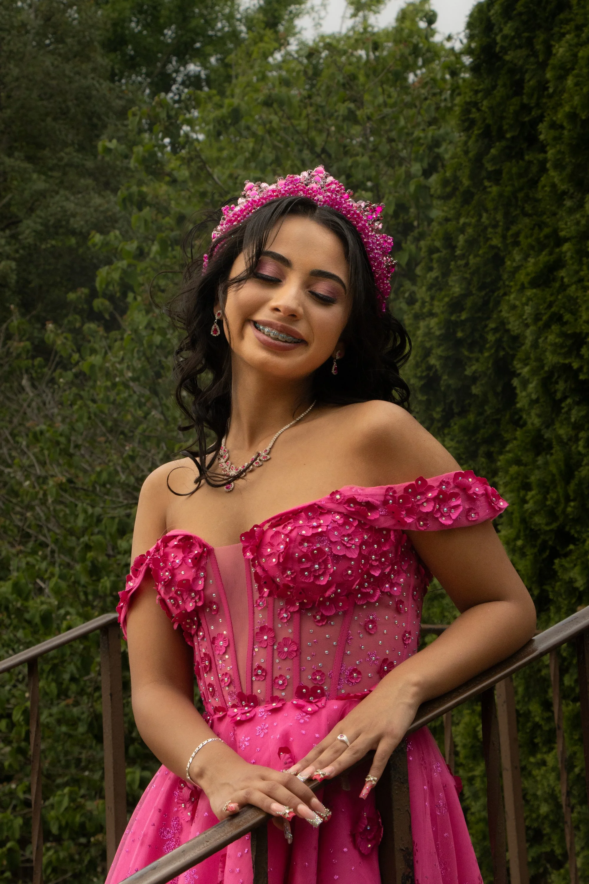 A young woman in a pink, floral, off-shoulder gown with beaded embellishments, standing outdoors on a curved wooden bridge, surrounded by green trees.