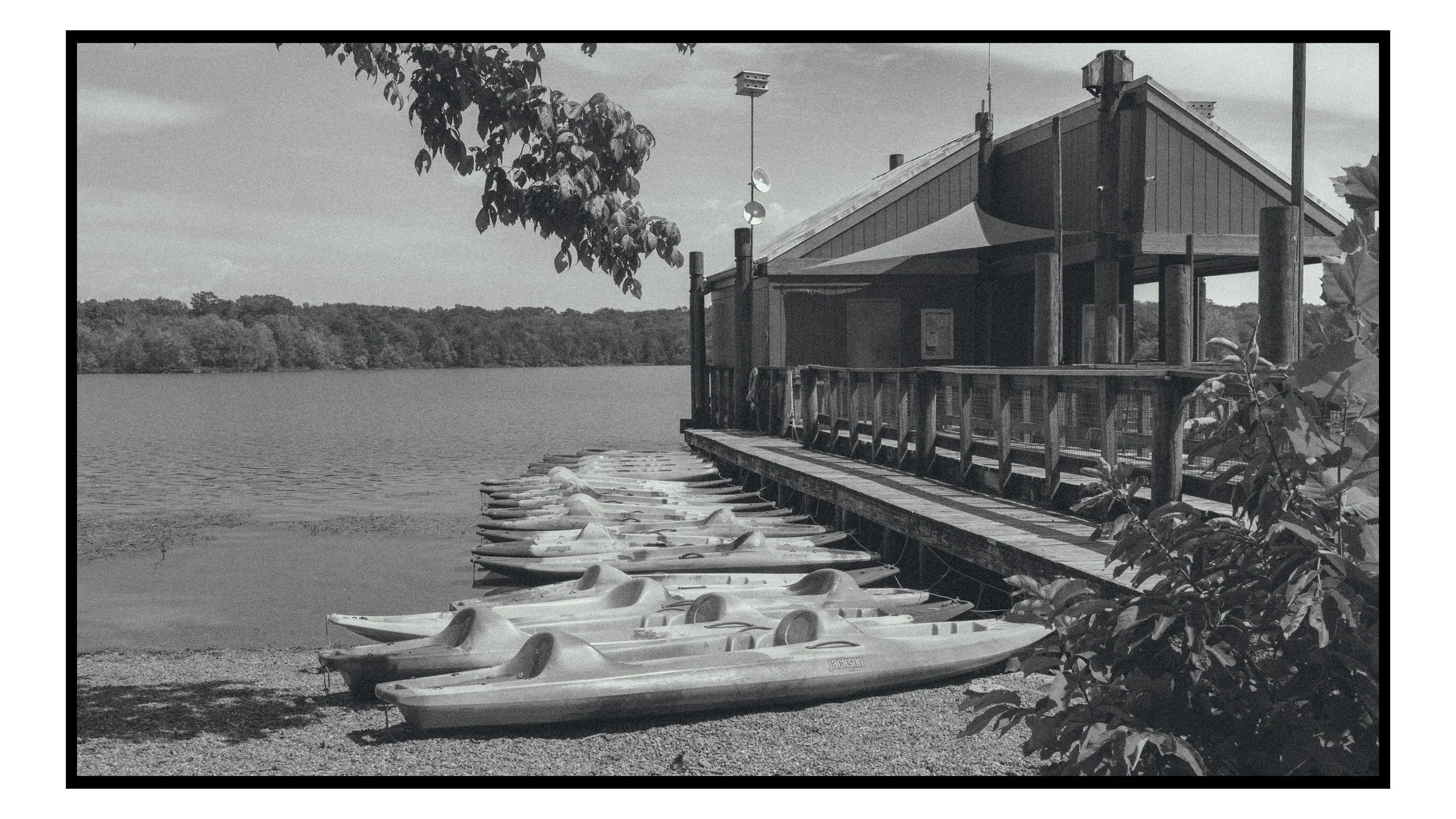 A lakeside scene in black and white, showing a row of kayaks or small boats lined up on the shore, a wooden pier with paddle boats and a lakeside building with a sloped roof, surrounded by trees and foliage.