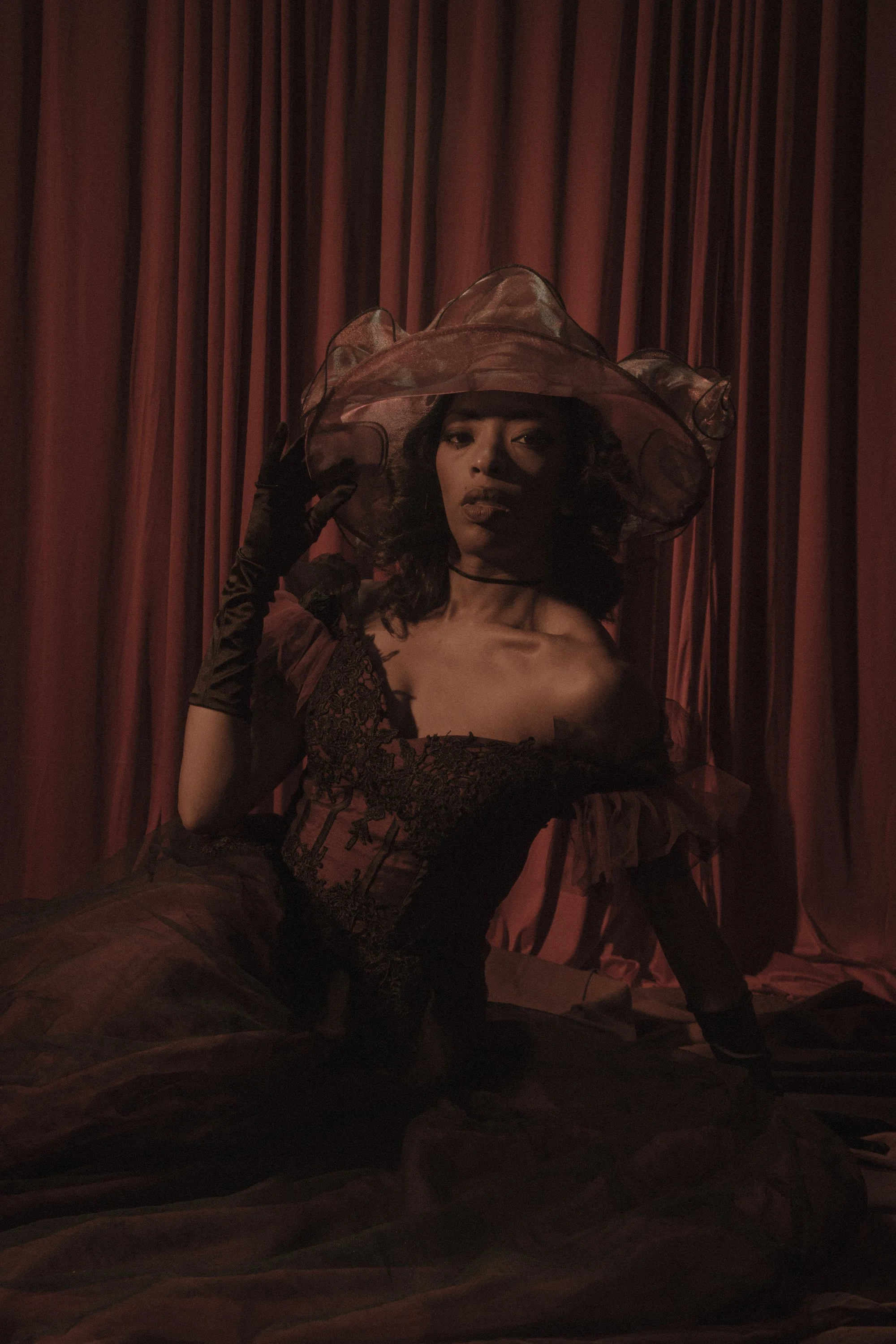 A woman dressed in vintage style clothing with a large hat, sitting on a chaise lounge against a red curtain background.