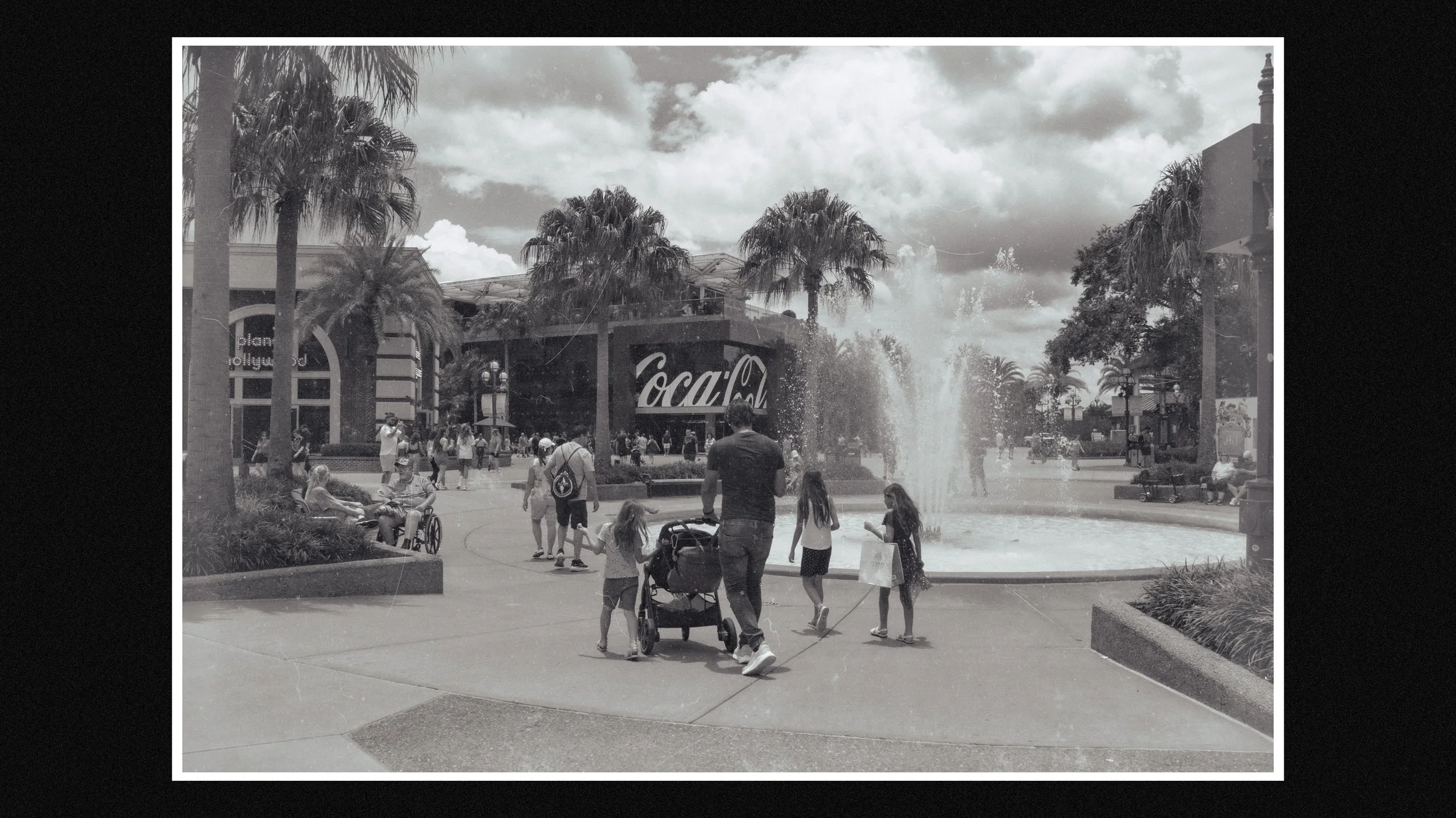 People enjoying time around a fountain at an outdoor shopping area with palm trees and large Coca-Cola signage.