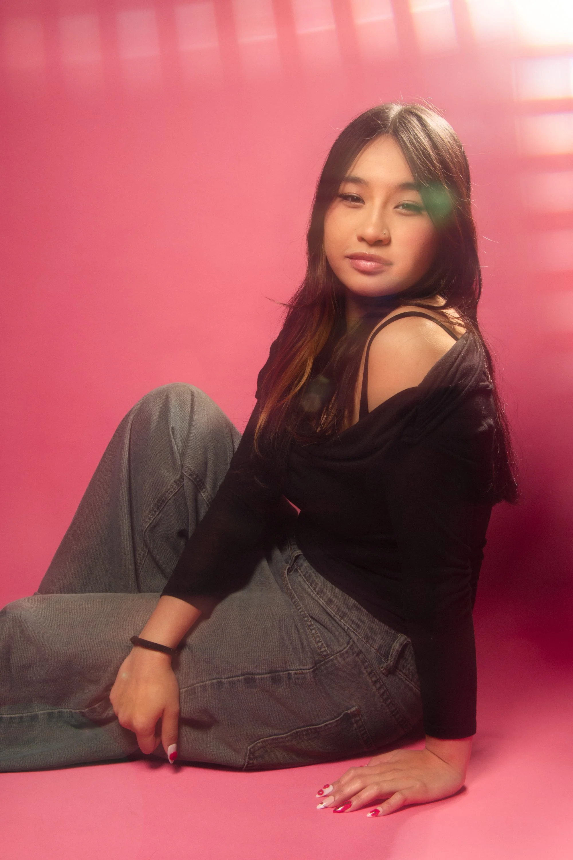A young woman with long dark hair sitting on a pink surface with a pink background, wearing a black top and gray jeans, looking at the camera with a slight smile.