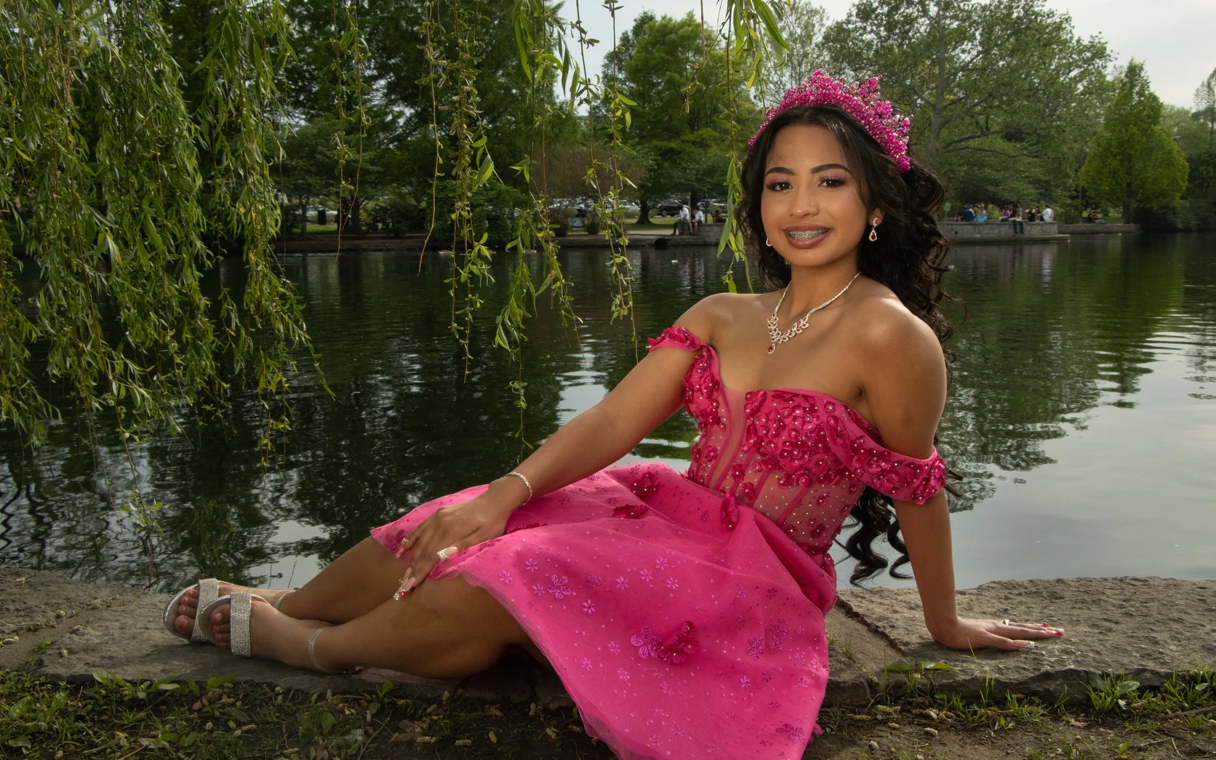 Young woman in a pink dress and tiara sitting by a river in a park during daytime.