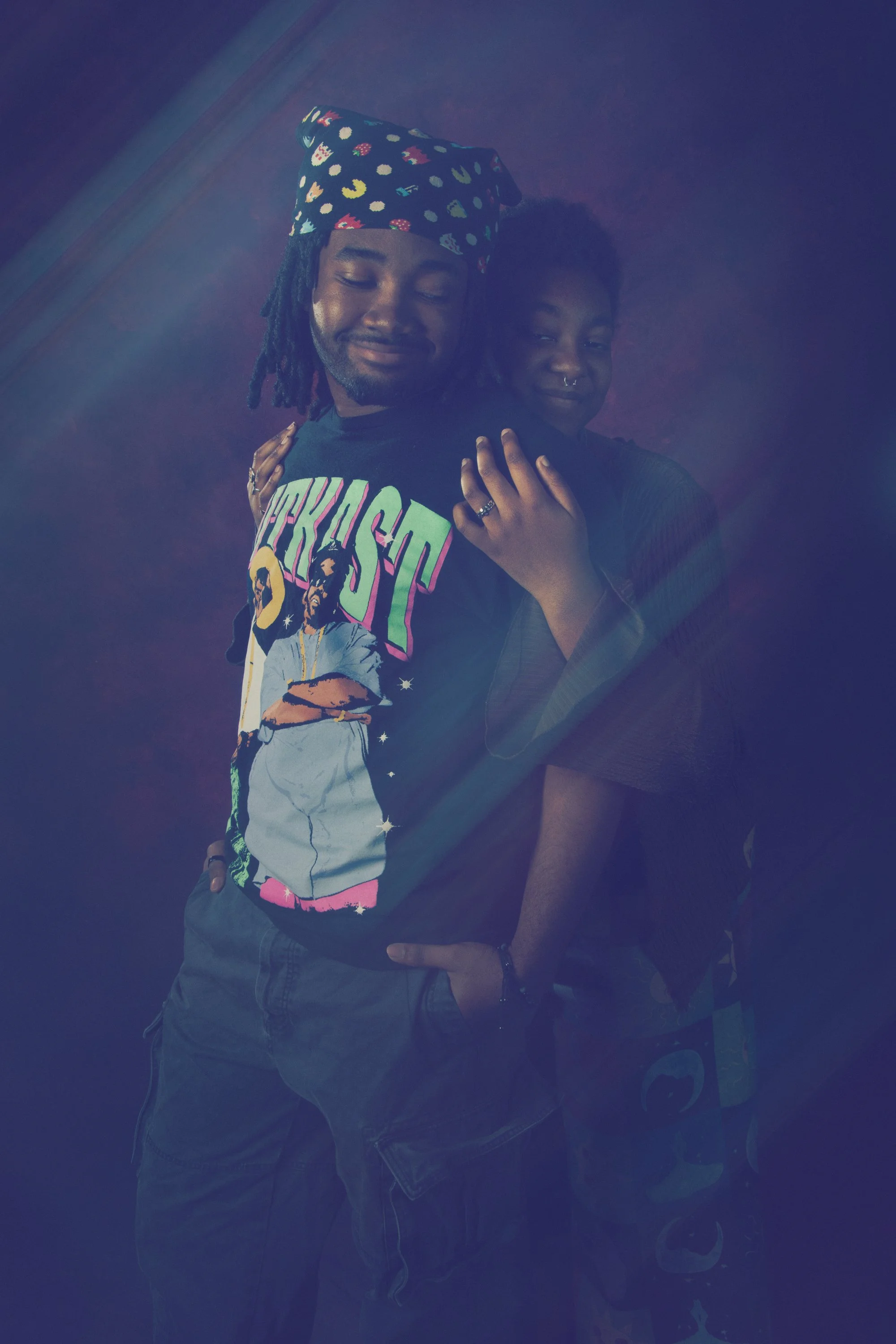 Two people posing together, one wearing a colorful graphic T-shirt and the other with a septum piercing and a dark top, both with their eyes closed and smiling softly.