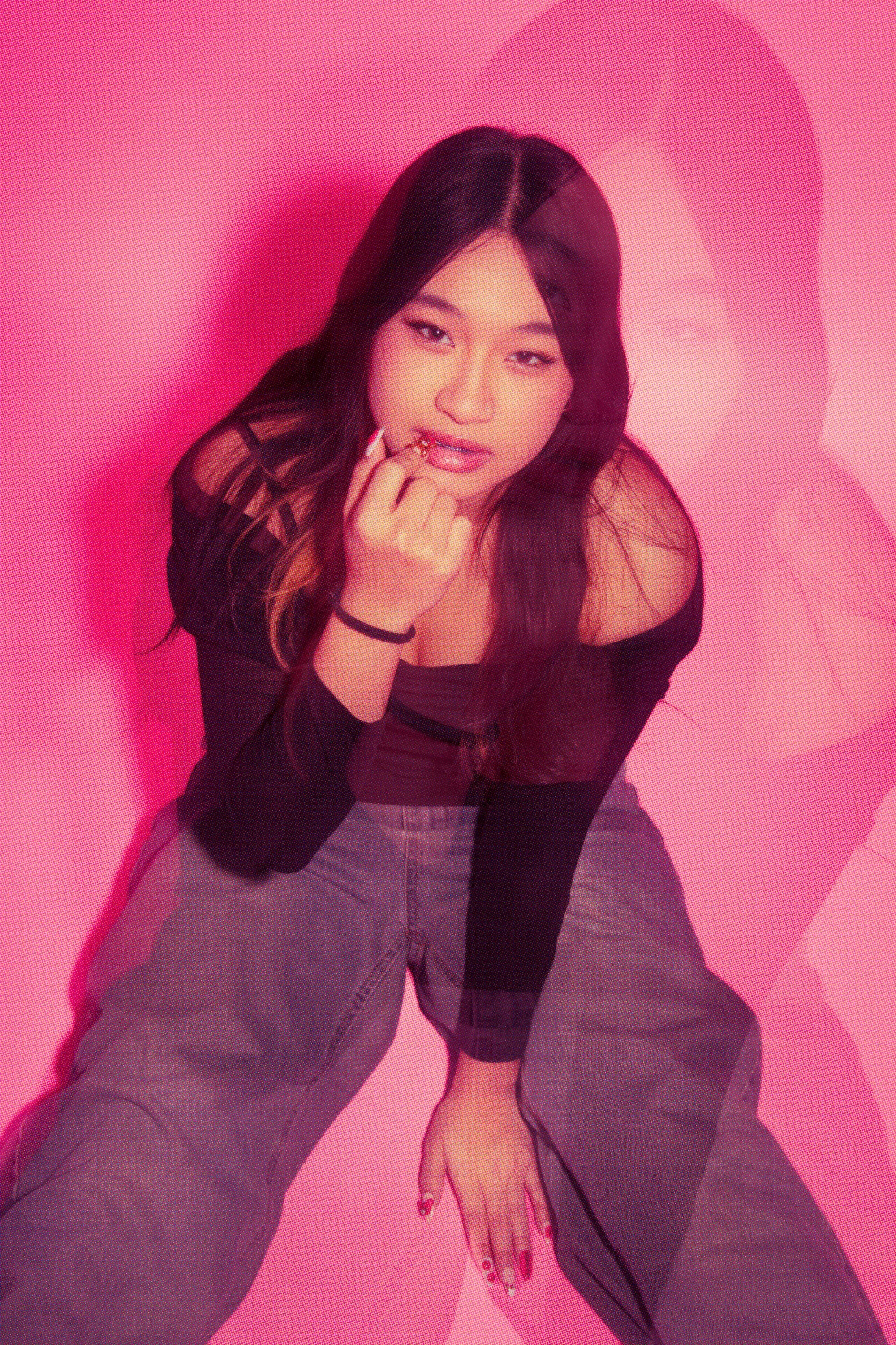 A woman with dark hair and light skin tone is sitting against a pink background. She is wearing a black top and gray pants, with her left hand resting on her knee. Her right hand is brought up to her mouth, and she is gazing directly at the camera wi