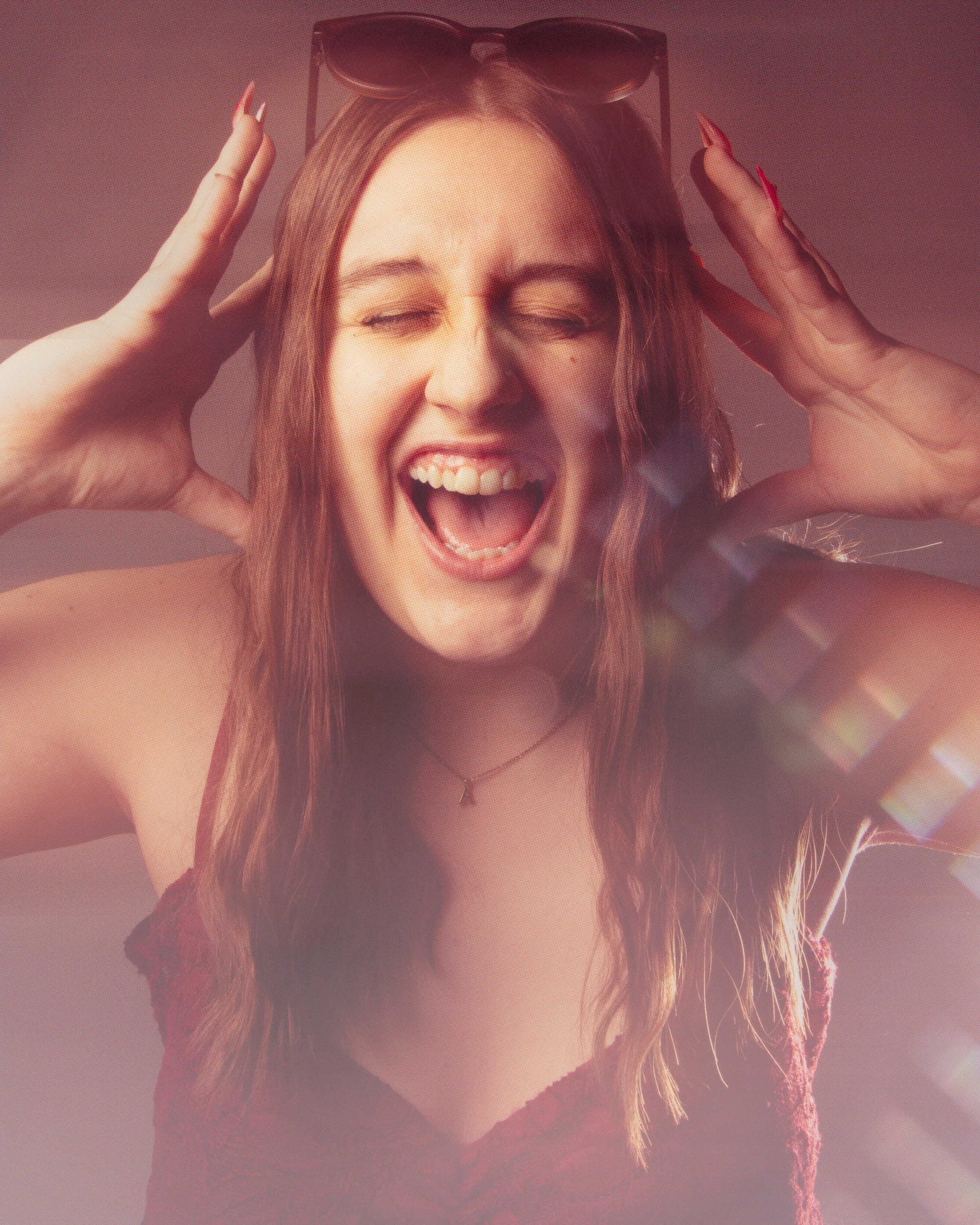 A young woman with long brown hair, wearing sunglasses on her head, is smiling with her eyes closed, hands on her head, wearing a red sleeveless top, and a necklace with a small pendant.